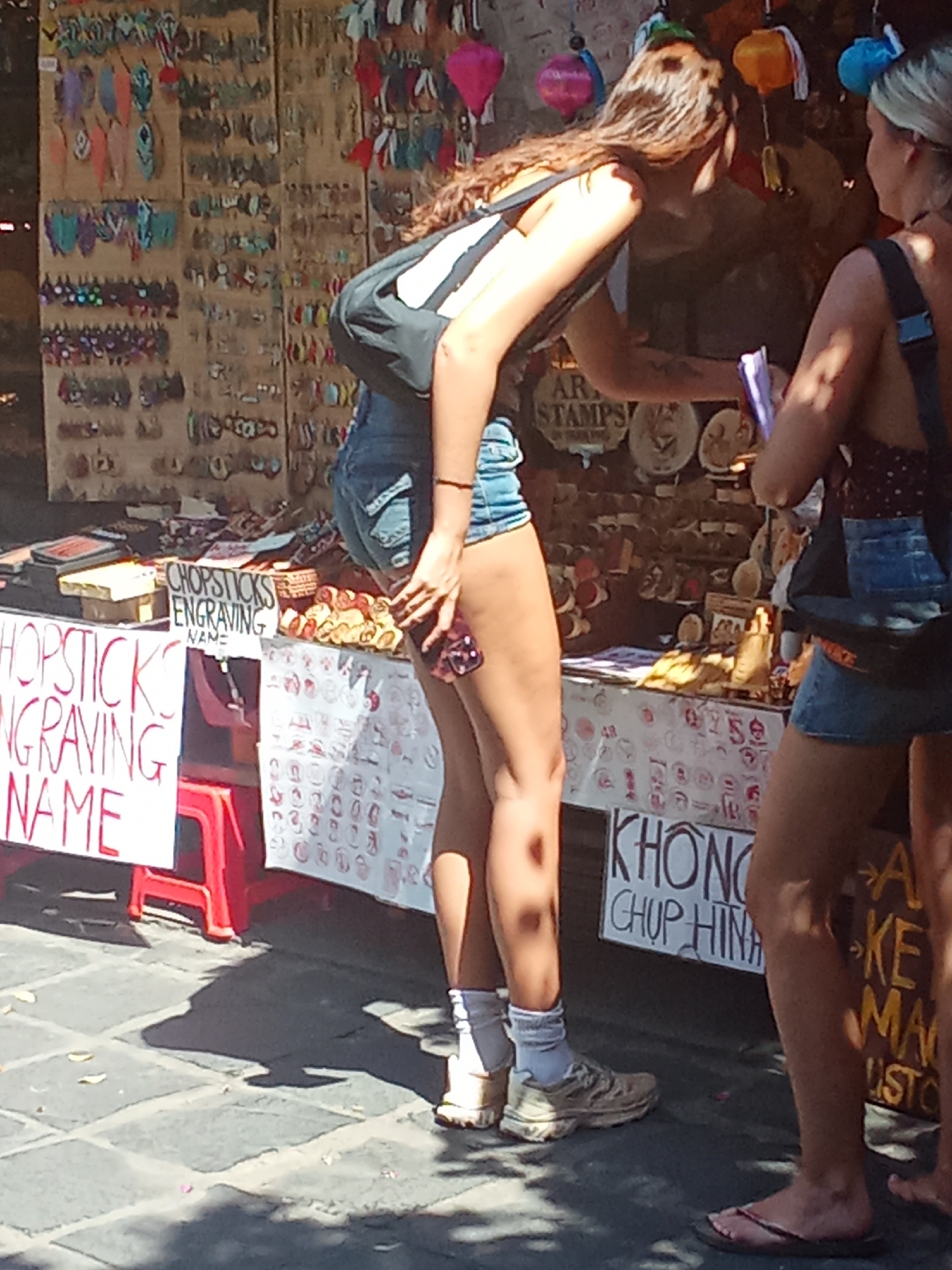 In Hội An Ward, Vietnam, two young women stand at an outdoor market stall. The woman on the left, with long brown hair, wears a black and white backpack and denim shorts. The woman on the right, with blonde hair, has a dark top with a patterned design and denim shorts. They appear to be browsing or interacting with the stall owner.
The stall displays a variety of items, including what appear to be decorative pieces, possibly souvenirs. There are also signs indicating services offered. One sign, written in red, reads "CHOPSTICKS ENGRAVING NAME". Another sign in the background has the text "AR STAMPS". Further down the stall, a sign reads "KHONG CHUP HINH" which translates to "No Photos". The market appears to be in full operation, with goods displayed on wooden shelves.
The scene suggests a daytime setting, possibly late morning or early afternoon, given the bright sunlight and shadows cast on the cobblestone ground. The atmosphere seems casual and typical of a bustling tourist market. The focus is on the people and the merchandise, highlighting a common tourist activity of shopping for souvenirs.