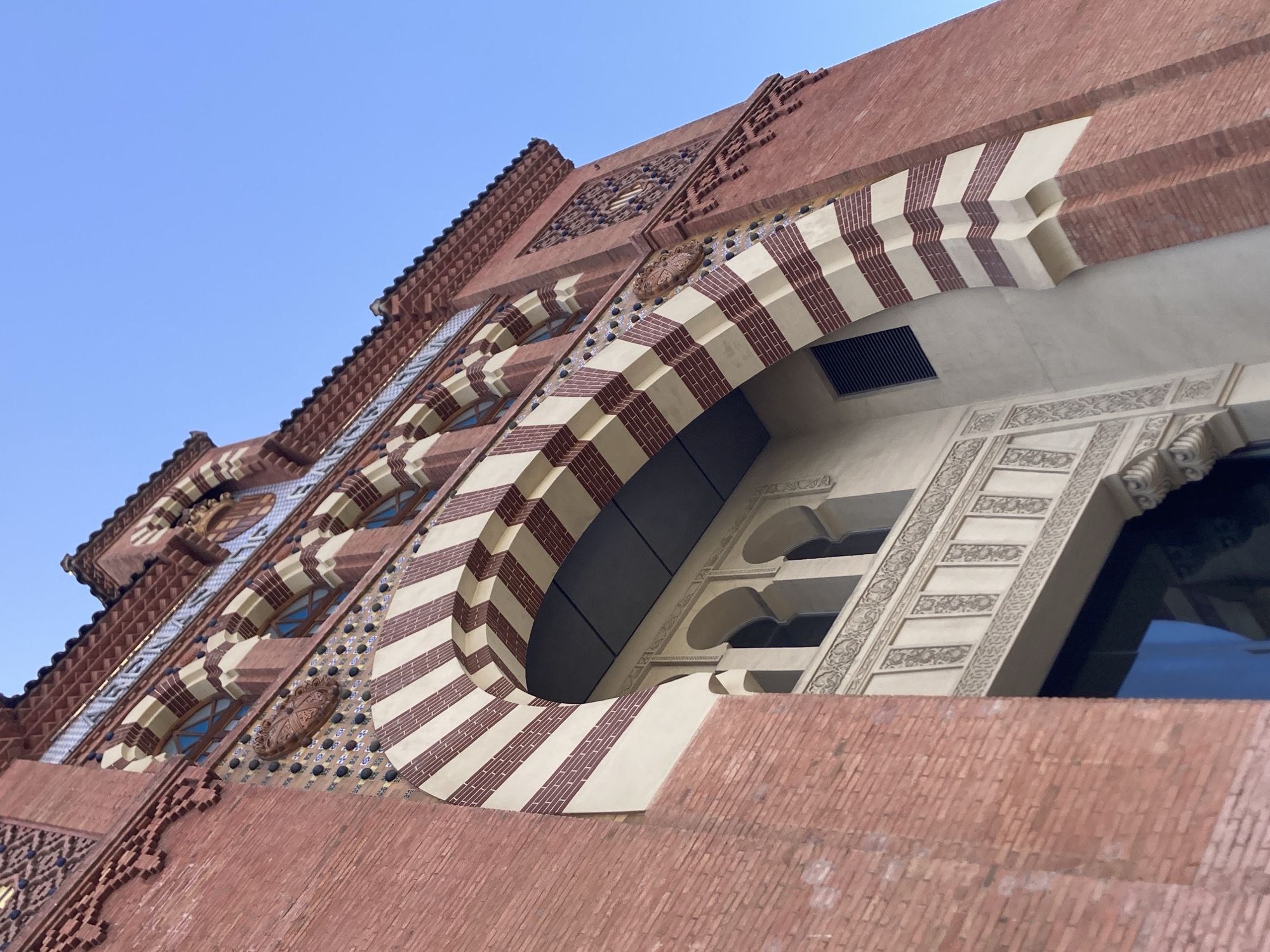 This is a low-angle, full-shot photograph of the exterior of the Mercat de la Boqueria in Barcelona, Spain. The market's facade is constructed primarily of red brick, with striking architectural details. A large, segmented arch, alternating between cream and reddish-brown stripes, dominates the lower portion of the image, framing an entryway. Above the arch, a band of decorative ceramic tiles featuring blue, brown, and cream geometric patterns is visible, interspersed with a circular, carved medallion. Further up, a series of arched windows with blue panes are set within red brickwork. Above these windows, a textured frieze and a tiled roofline are apparent. The sky above is a clear, bright blue. The overall style of the architecture suggests Moorish influences, particularly in the geometric patterns and archways. No people are visible in the frame, and the scene is static. The image focuses on the architectural grandeur of the building's facade.