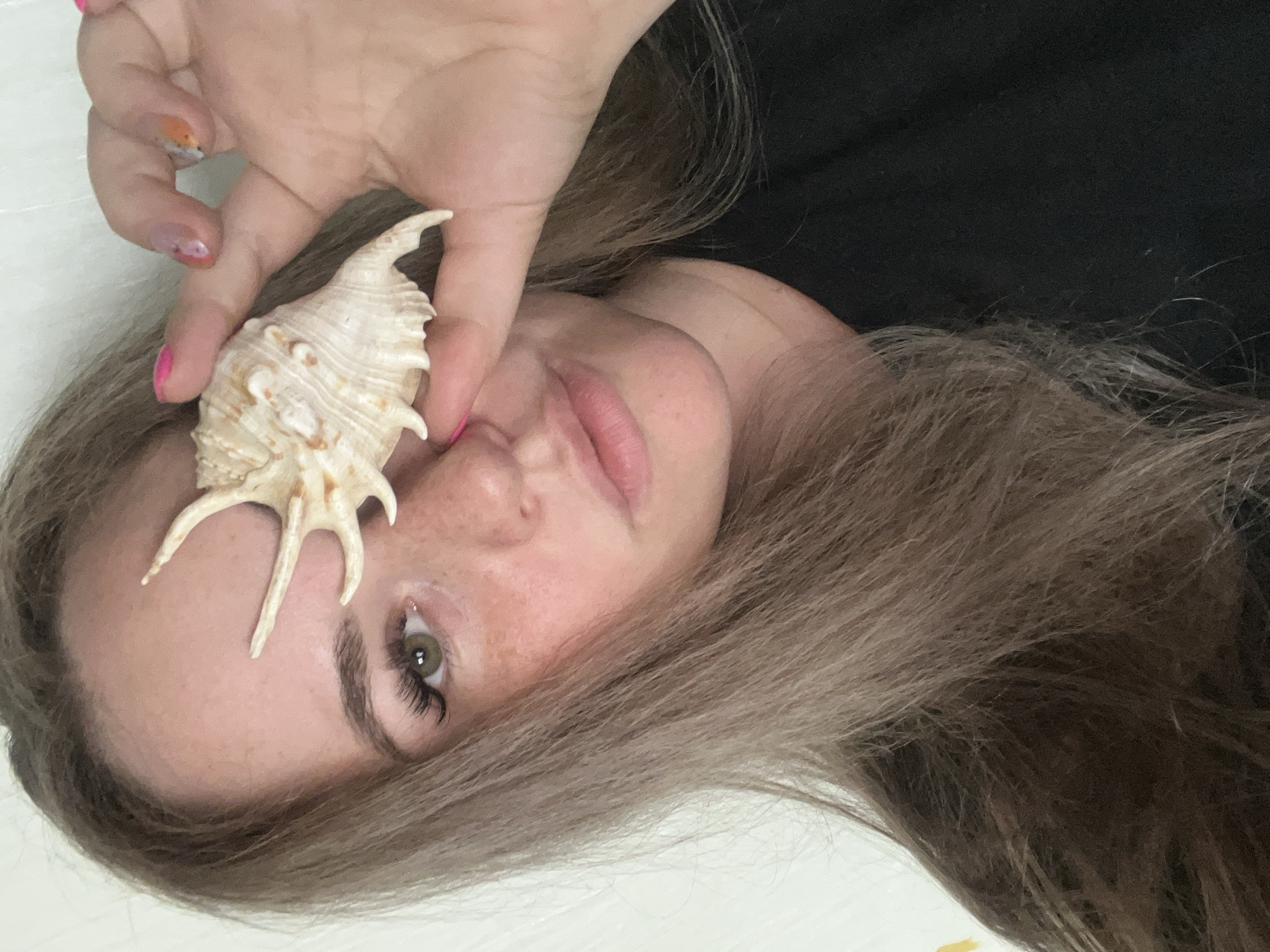 A person is holding a seashell in front of their face. The person has light brown hair, freckles, and green eyes with long eyelashes. The person's hand is visible holding the seashell, with some pink nail polish on their nails. The background is a mix of black fabric and a white surface. The city is not visible and therefore, its location is unknown.