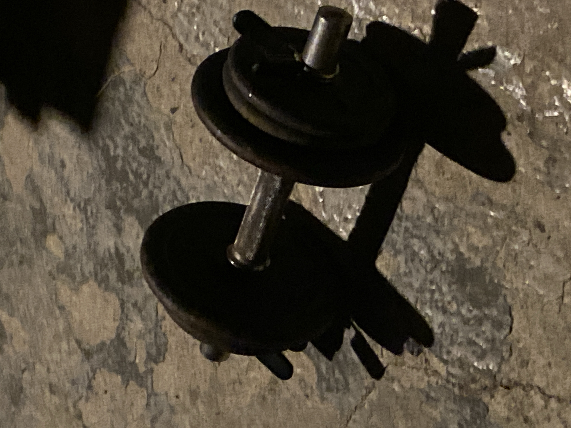 A dark, angled, close-up shot features a dumbbell with two weight plates attached to each side. The dumbbell and its shadow are primarily black. The dumbbell appears to be resting on a rough, textured, gray surface, which could be concrete or stone. The lighting is dim, casting long shadows. The image appears to be taken indoors. There is no indication of the image being taken in the city of Konstantinovo, Bulgaria, or the presence of people or interactions.