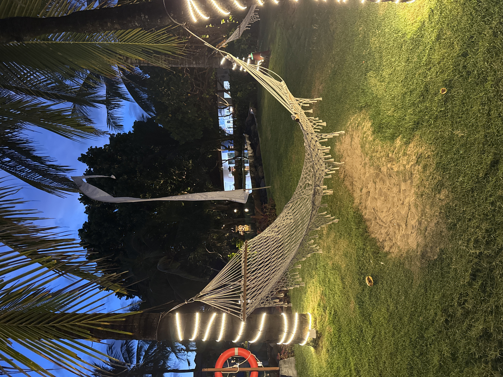The image depicts an outdoor setting in Kuta Selatan, Indonesia, at night. A white rope hammock is strung between two palm trees, illuminated by string lights. A red life preserver is visible near one of the trees. The ground is covered in green grass, with a patch of sand near the hammock. The sky is a deep blue, visible through the palm fronds. A building is visible in the background, illuminated by lights. There are no people visible in the image.