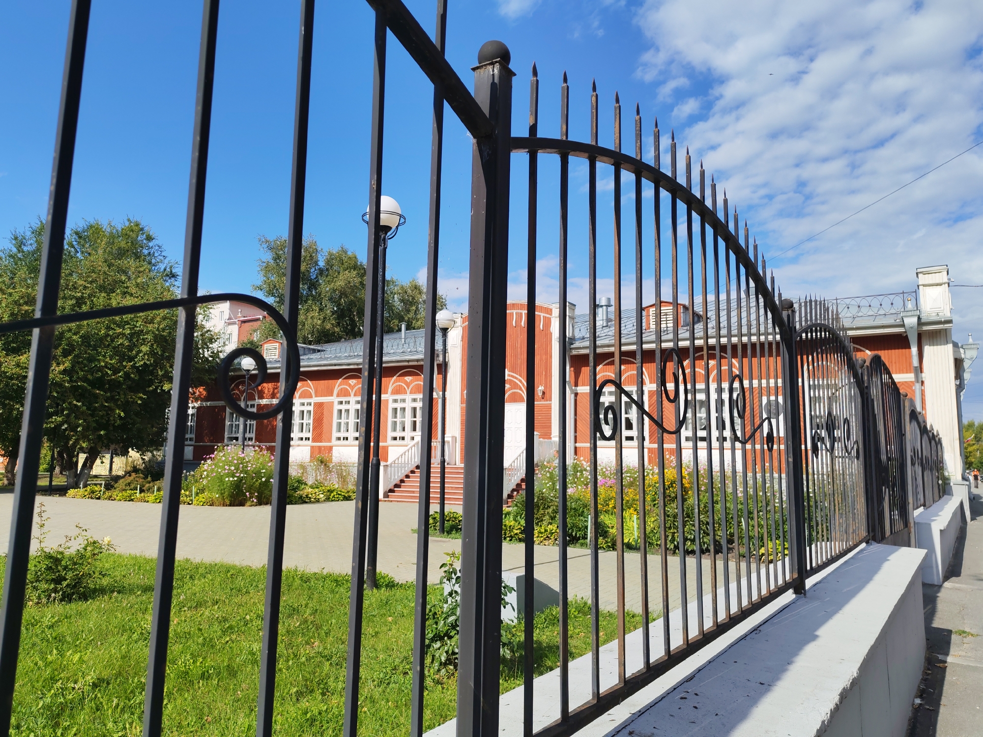 A medium shot captures a wrought iron fence in the foreground, with its pointed finials visible against a bright blue sky dotted with white clouds. Behind the fence, a red brick building with white trim and arched windows stands prominently. Green grass and a paved walkway are visible in the middle ground, with trees and flowering bushes adding color to the scene. The building appears to be a historical or public institution, given its architectural style and well-maintained grounds. The overall atmosphere is bright and sunny, suggesting a pleasant day outdoors.