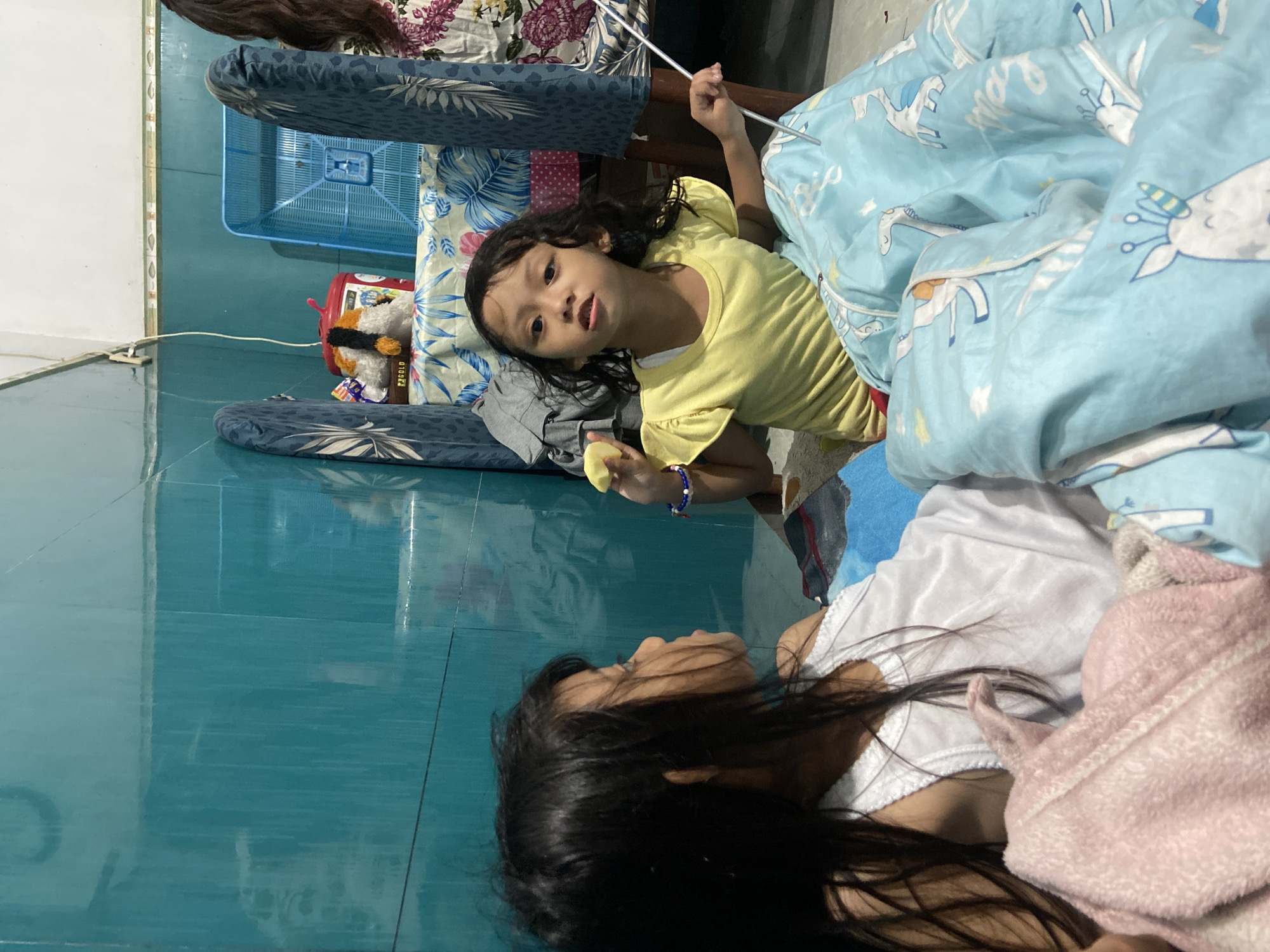 Two young girls are positioned on the floor, likely in Gelam, Indonesia, as indicated by image geotags. One girl wears a yellow shirt and holds a light yellow object (possibly food). She looks toward the camera and holds a white stick in her other hand. She is partially covered by a light blue unicorn-patterned blanket. A variety of objects surround her, including a patterned chair, a blue basket, and toys. The other girl wears a white shirt and is partially covered by a pink towel and the blue blanket. She looks at the girl in the yellow shirt. The floor is tiled in turquoise.