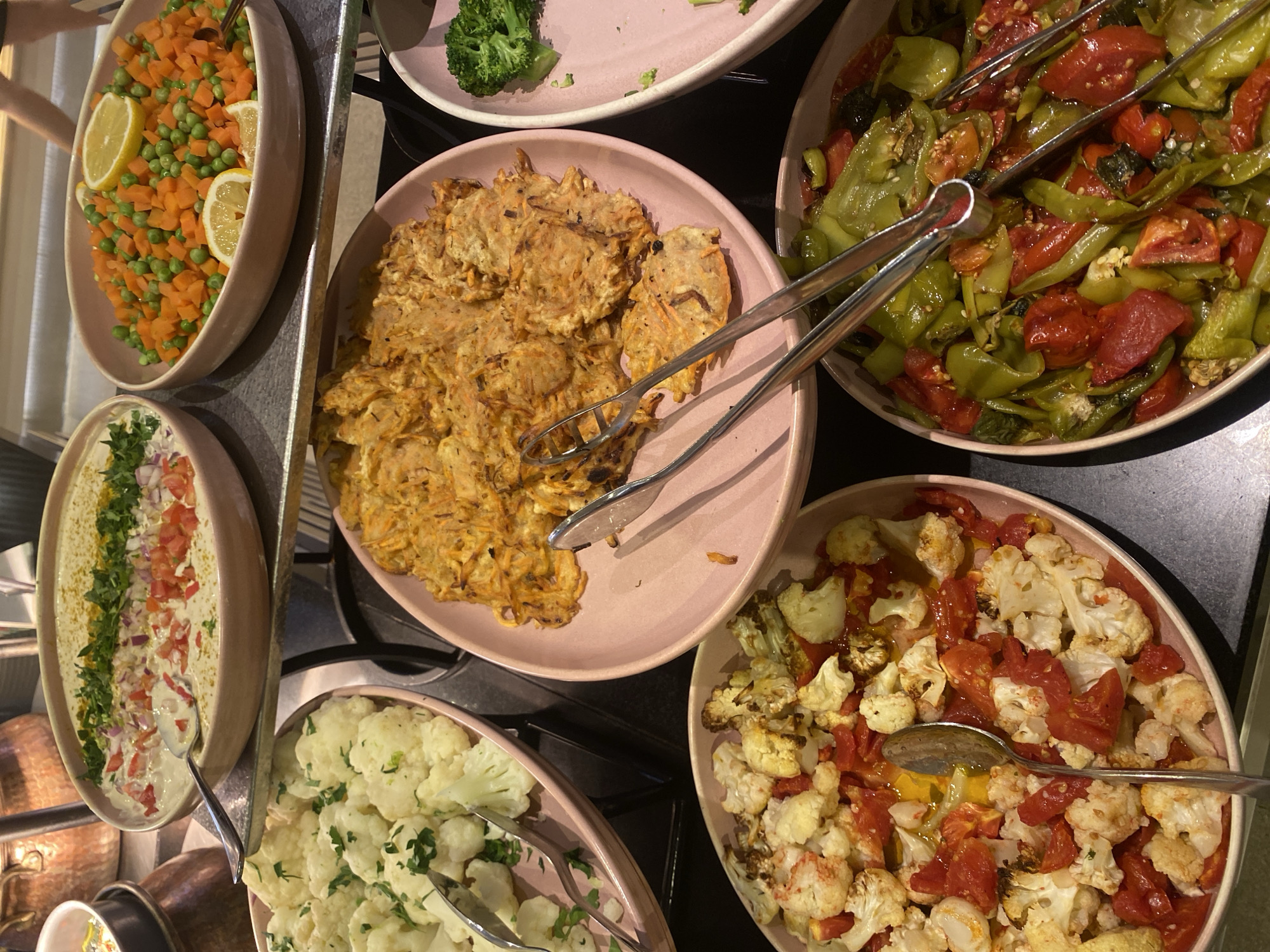 In Aksu, Turkey, a buffet-style display of various dishes is presented. The foreground features a close-up of a pink oval dish containing roasted cauliflower florets mixed with chopped red tomatoes and a glistening yellow oil. A silver spoon rests within this dish. To the right, another pink oval dish is filled with a colorful medley of sliced green peppers and red tomatoes, with serving tongs resting on top. Behind these, a light pink oval dish holds what appears to be grated vegetable fritters, possibly made from carrots, and serving tongs are positioned over them. Further back, a larger pink oval bowl contains a mixture of diced carrots and green peas, garnished with lemon slices. To the left, a large, shallow bowl, possibly ceramic, is filled with a creamy white dip, topped with finely diced red tomatoes, red onions, and fresh green herbs, with a spoon inserted. The dishes are arranged on a dark gray surface, with a glimpse of copper pots in the background, suggesting a restaurant or catering setting. The lighting is warm and consistent, illuminating the food items clearly.