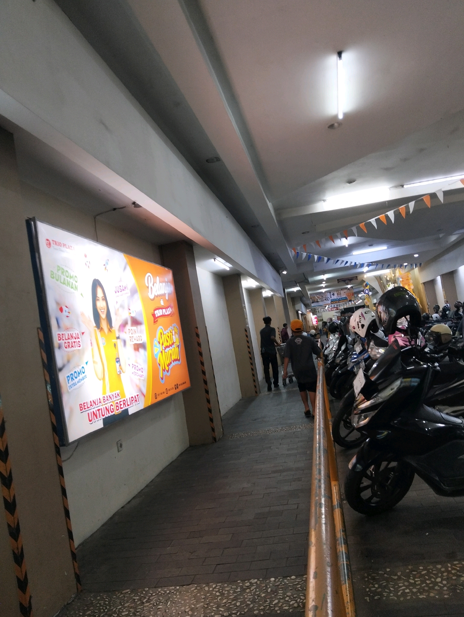 The image depicts a covered outdoor area, likely a parking lot or walkway, in what appears to be a commercial district. The ceiling features fluorescent lighting and decorative triangular flags in orange, white, and blue. On the left, a large illuminated billboard displays an advertisement for "TRIO PLAZA," featuring a woman in a yellow shirt promoting "PROMO BULANAN" (monthly promo), "BELANJA GRATIS" (free shopping), and "PROMO EXTRA MEMBER." Further down the walkway, several motorcycles are parked, with some individuals walking and others attending to the vehicles. The ground is paved with dark rectangular tiles and interspersed with lighter speckled sections. The overall environment suggests a retail or service area in an urban setting, possibly a shopping center or a market. The presence of the billboard and parked motorcycles points to a commercial and public space. Given the context of Trio Plaza being a known shopping mall in Magelang, Indonesia, this image is likely taken within its vicinity.