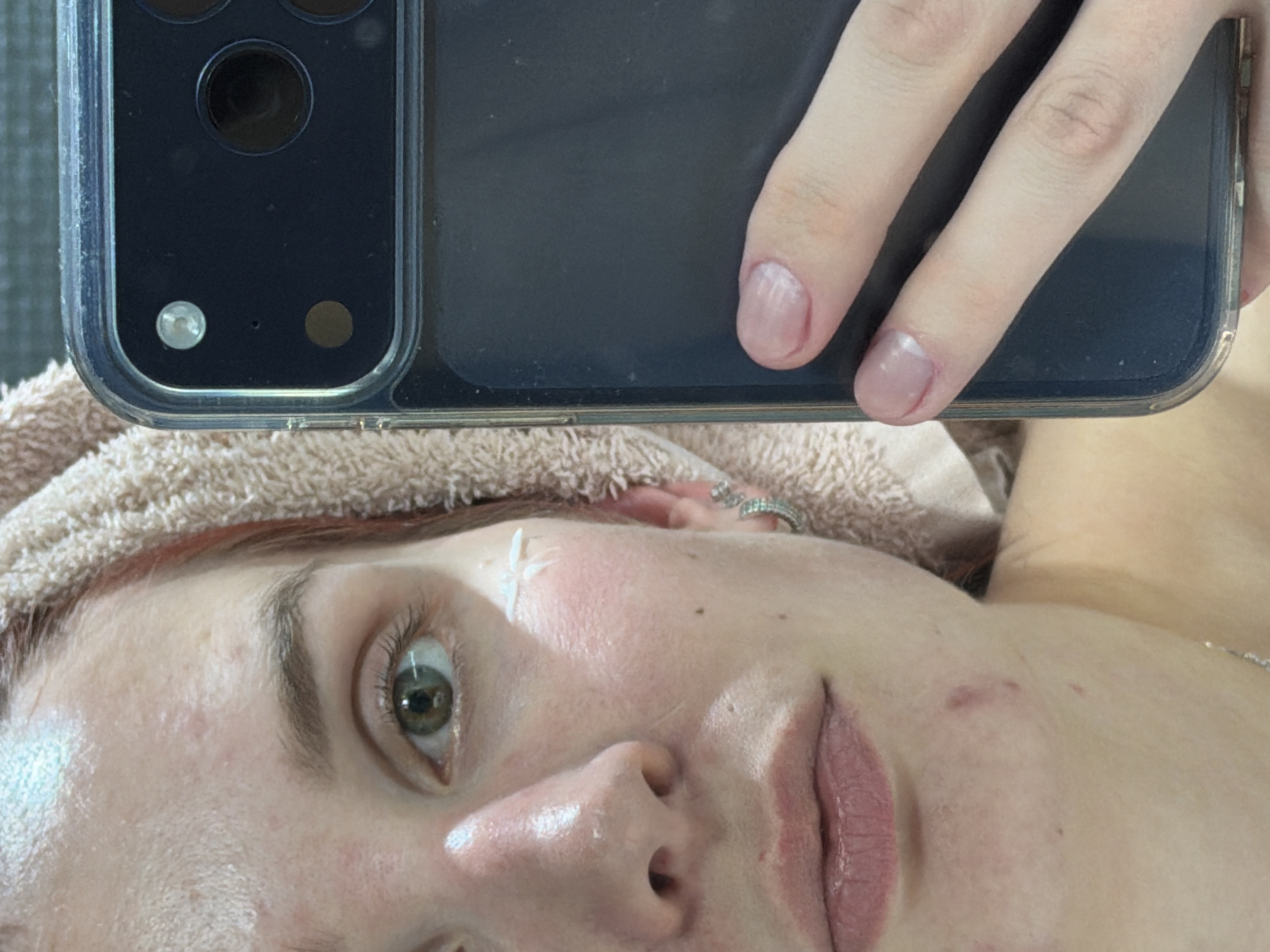 A selfie taken from above focuses on the face of a fair-skinned person with a towel wrapped around their head. A smartphone, presumably used to take the picture, obscures the upper portion of the scene, with fingers visible holding the device. The person's face features a green eye and a smear of white cream on their cheek. The background is not readily discernible, offering no clues regarding the location in Chaikovsky, Russia, or any specific environmental details. No specific actions or interactions can be inferred beyond the act of taking the selfie.