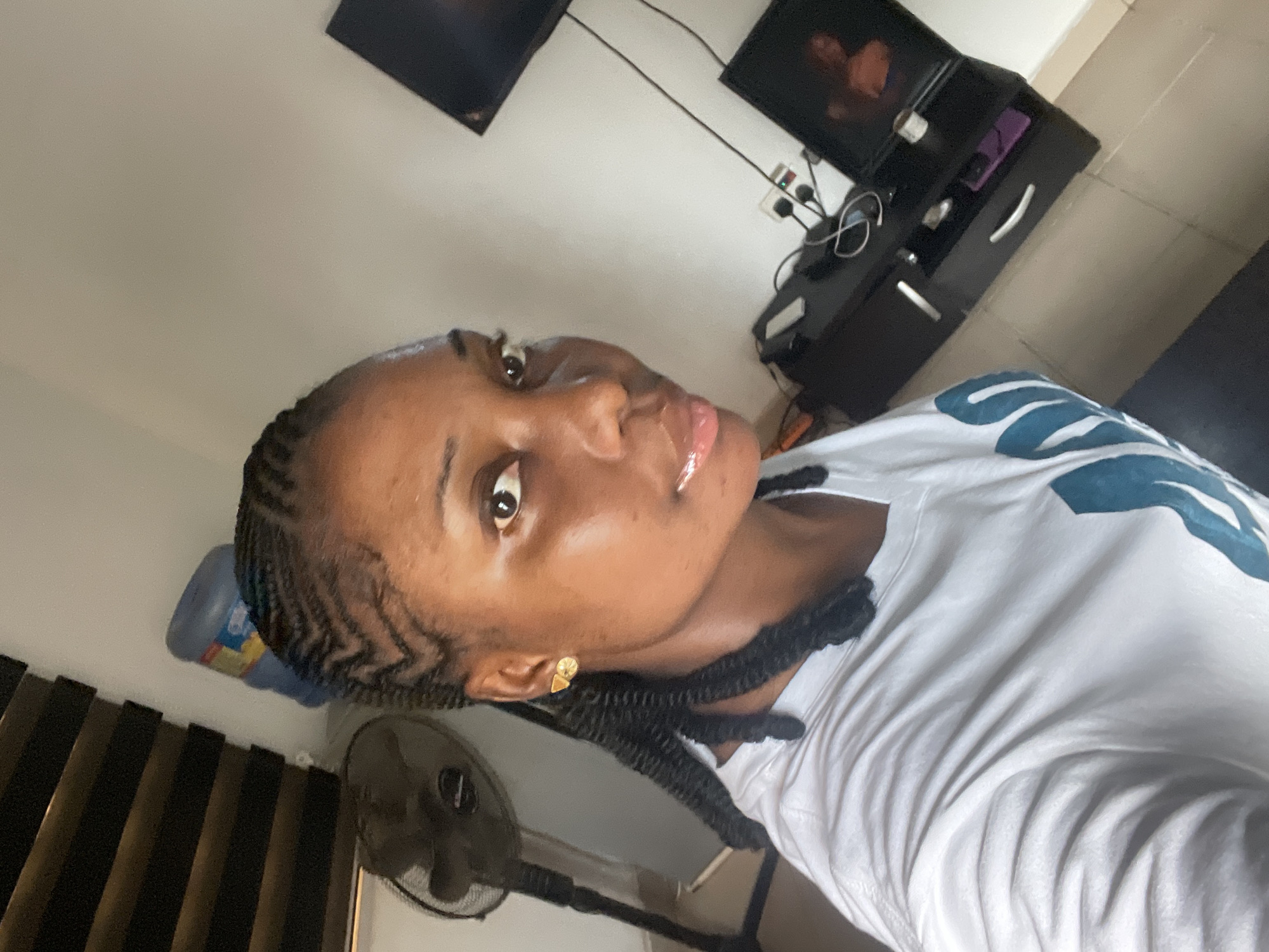 A selfie captures a young Black woman indoors, likely in Abuja, Nigeria, with a calm and poised expression, looking directly at the camera. She has intricate cornrow braids styled across her scalp, leading into longer individual braids that fall to her shoulders. She wears a white t-shirt featuring a large blue and white abstract design on the right side. Small, gold-toned stud earrings are visible on her left ear.
The setting appears to be a residential room, possibly a living room or bedroom. In the background, to the right, is a black entertainment unit or TV stand. Above it, two flat-screen televisions are mounted on a light-colored wall, both displaying faint, indistinct images. Electrical wires and a white power strip are visible behind the stand. To the left, a large blue water dispenser bottle rests on a surface, and below it, a black standing fan is positioned. The room features light-colored walls and tiled flooring. The overall lighting suggests it is daytime or a well-lit indoor environment. The only visible text, on the blue water bottle, is not clearly legible.