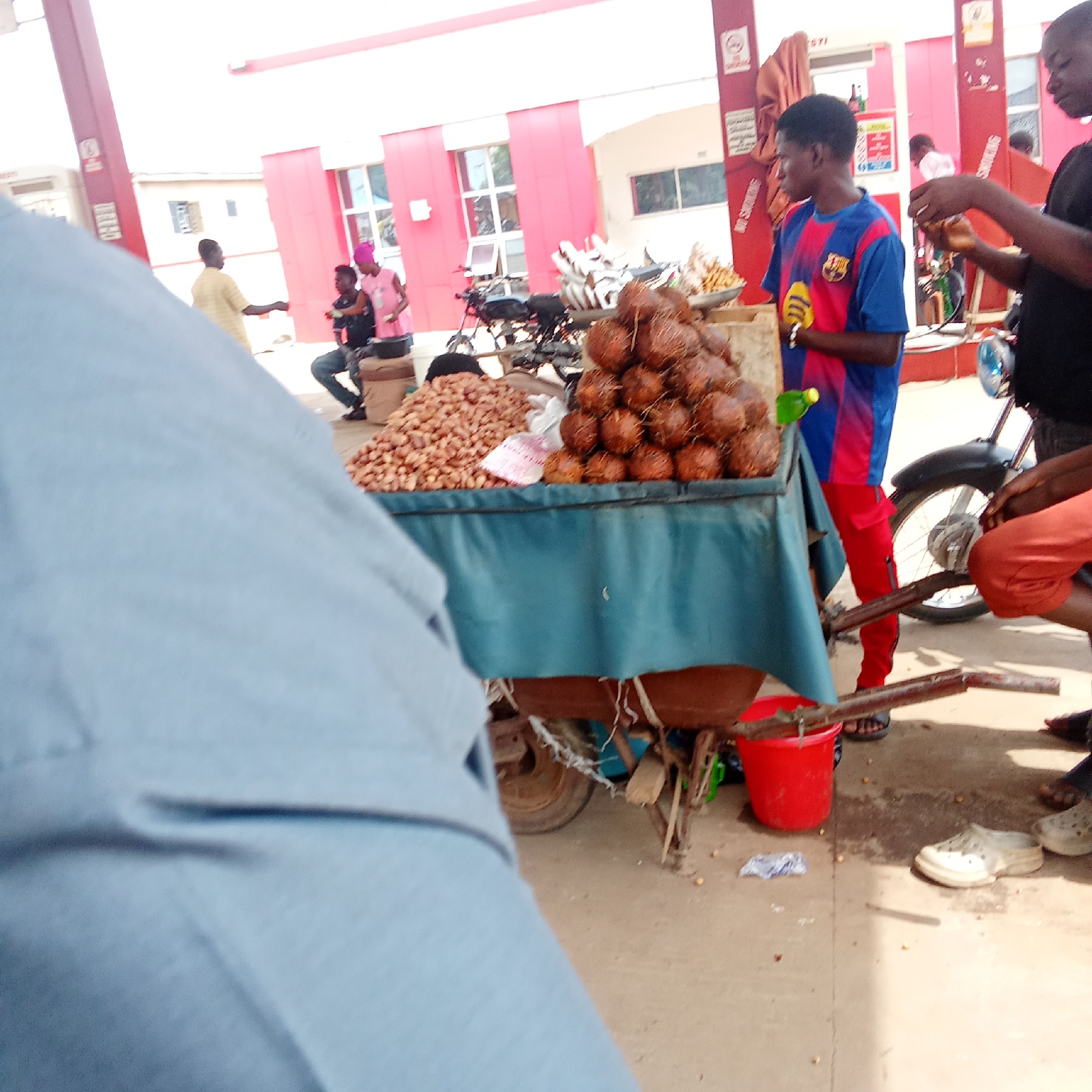 The image depicts a street scene, possibly a roadside market, in an urban area. The focal point is a cart laden with peanuts and a stack of coconuts, covered with a blue cloth. Several people are visible, including a vendor wearing a blue and red Barcelona jersey. Other individuals are standing or sitting nearby, some seemingly interacting with the vendor.
In the background, there's a building with red and white accents, possibly a gas station or a commercial building, with visible signs. A motorcycle is parked beside the cart, and a red bucket is placed near the cart's wheel. The setting appears to be a paved area.