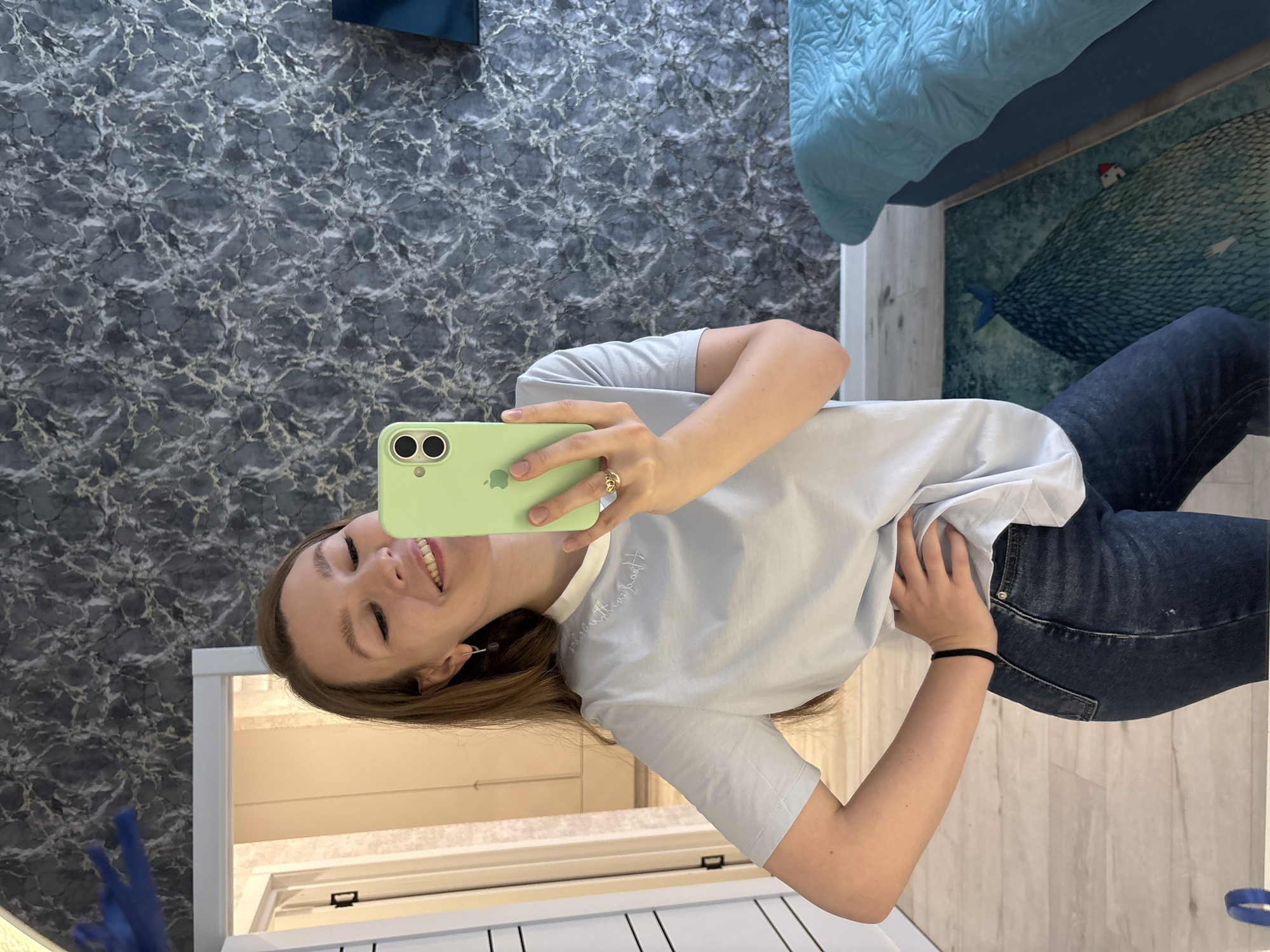 The image shows a young woman taking a mirror selfie in a room. She is wearing a light blue t-shirt and dark blue jeans, holding a light green smartphone in her right hand. She has long brown hair and is smiling at her reflection. Her left hand is on her hip. The background of the room features a grey and white marbled wallpaper and a partially visible blue bed with a quilted comforter. To the left of the woman, a doorway leads to another room with light-colored walls and doors. On the floor, there is a rug with a dark green pattern depicting a large, textured whale-like shape with a small red-roofed house and a figure. The overall scene suggests an indoor, residential setting. There is no information to suggest this location is in Izhevsk, Russia.
