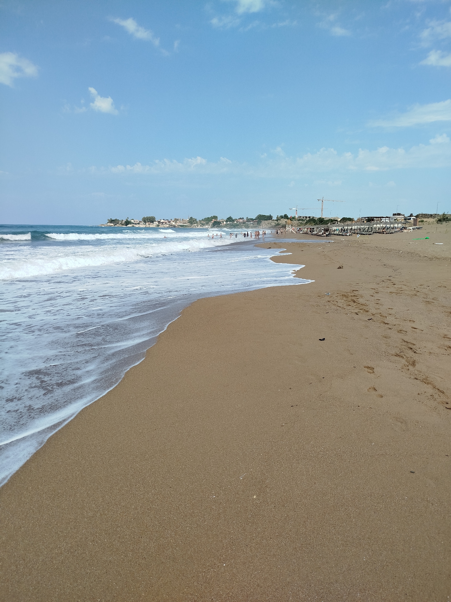 A wide, sandy beach stretches along the coast with gentle waves lapping at the shore. In the distance, a line of buildings and construction cranes suggest a developing urban area. A few scattered footprints mark the sand, indicating recent human presence. The sky above is a clear blue with a few wispy clouds. The scene is likely located in Manavgat, Turkey, known for its coastal resorts and ongoing development.