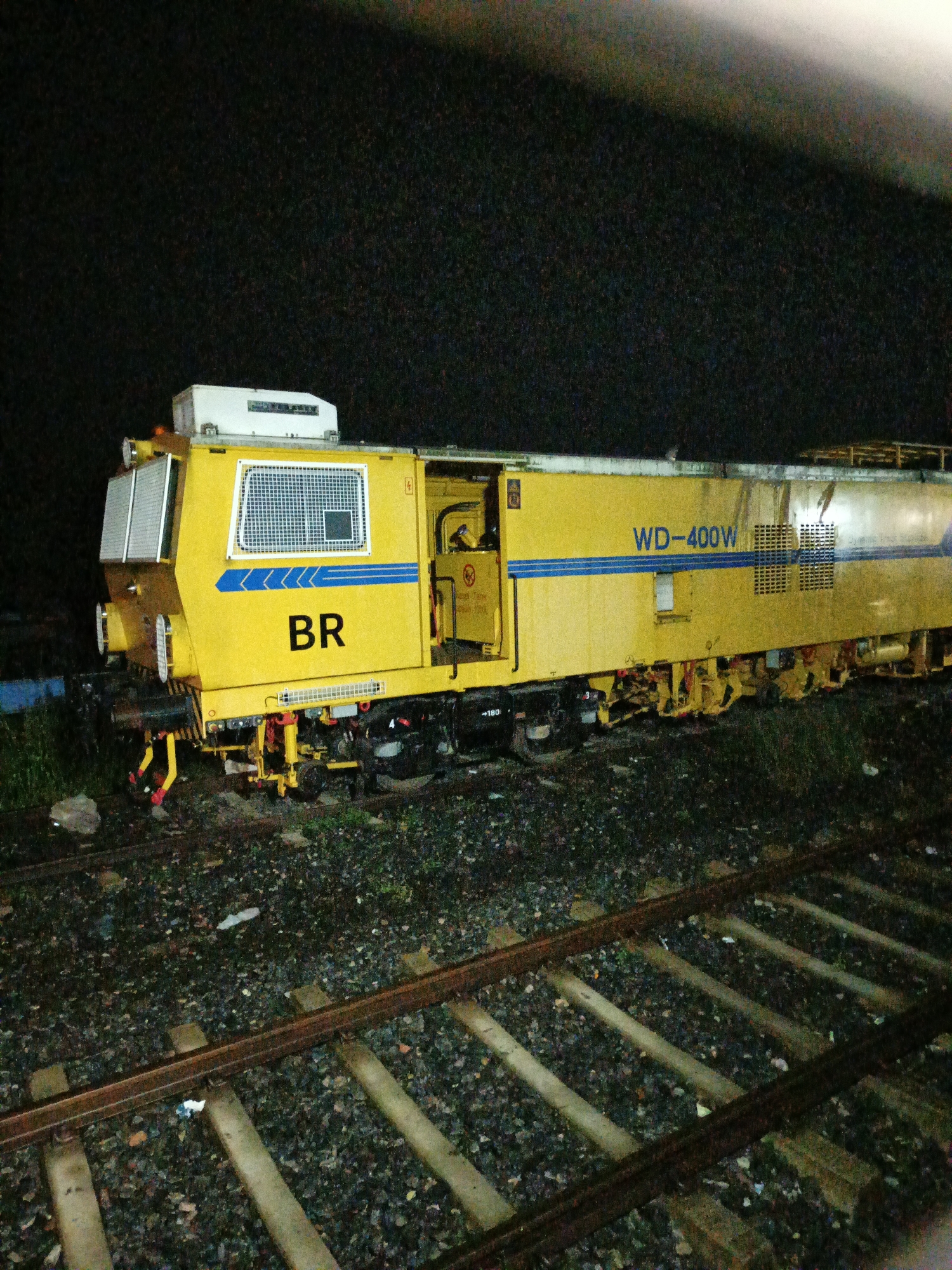 An eye-level, medium shot captures a yellow railway maintenance vehicle, identified by the markings "WD-400W" and "BR," stationary on railway tracks at night in Rajbari, Bangladesh. The vehicle, a prominent yellow color with blue accents and a white upper section, occupies the majority of the frame. Its complex machinery, including various levers, wheels, and a lowered set of tools, is visible beneath its body. The foreground consists of ballast and several sets of parallel railway tracks, with some scattered debris. The background is dark, suggesting nighttime, with a slightly brighter area at the top, possibly from artificial lighting or a distant light source. The scene appears to be an industrial or operational area related to railway maintenance. No individuals are visible in the image.