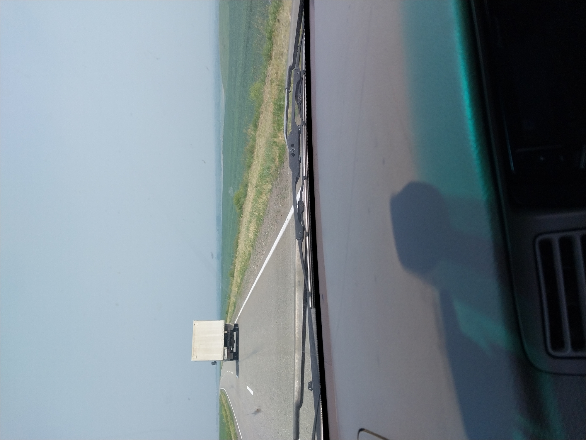 A white box truck is seen from a vehicle's perspective, driving on a paved road. The truck is traveling away from the viewer, in the same direction as the vehicle. The road is divided by a solid white line on the left and a dashed white line on the right. The environment is rural, with green fields on either side of the road stretching to the horizon. The sky is a pale blue and appears hazy. The image is captured through the windshield of the vehicle, with a portion of the dashboard and windshield wipers visible in the foreground. The license plate on the truck appears to read "R449TA".
