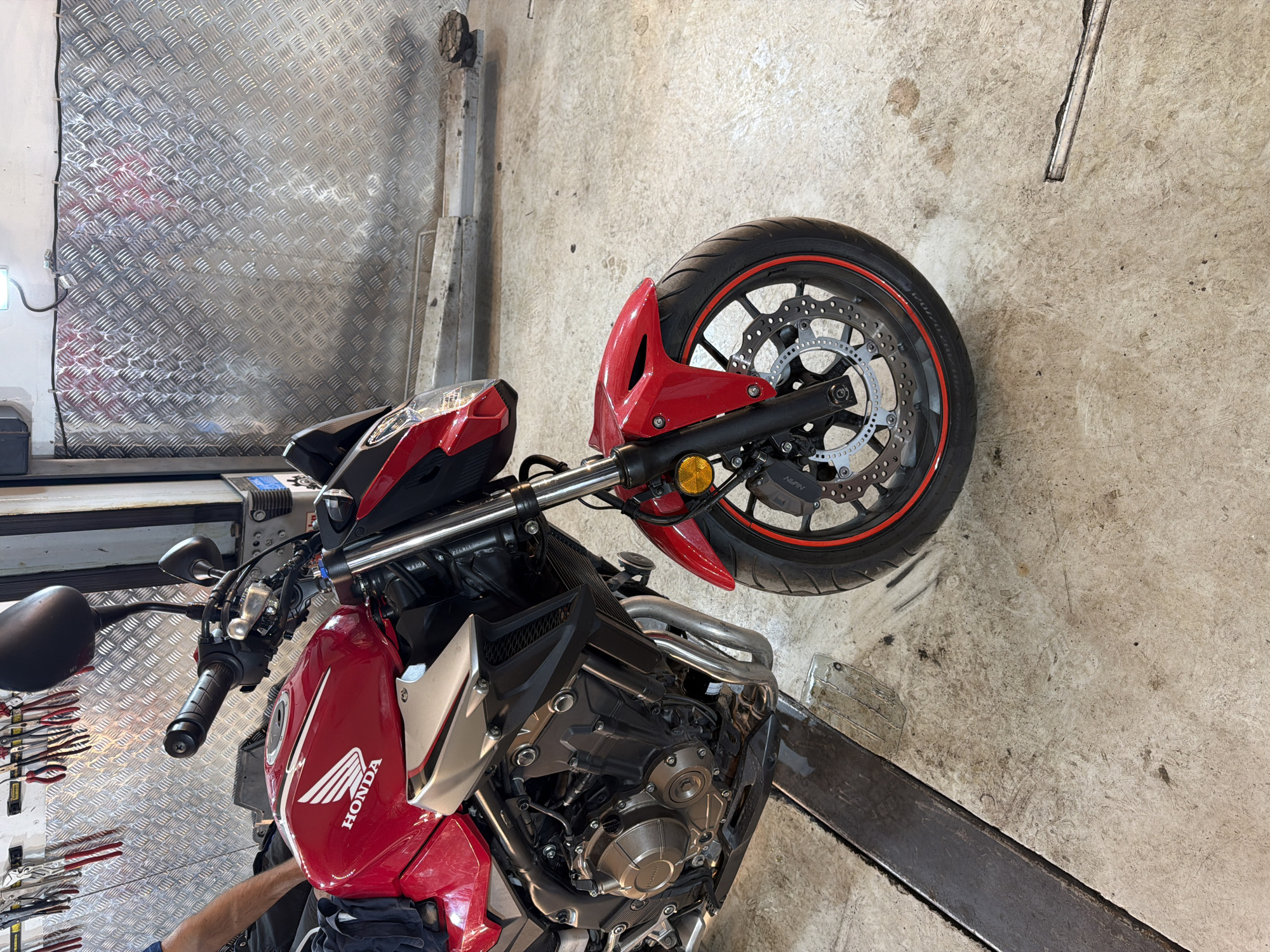 The image shows a red and black Honda motorcycle in a garage. The motorcycle is parked on a lift, with its front wheel visible in the upper right corner of the frame. The motorcycle has a "HONDA" logo on the red section of the body. A tool rack with various tools is visible on the left side of the image. The background consists of a concrete floor and a diamond-plated metal wall. The exact location in Sofia, Bulgaria cannot be determined from the image.