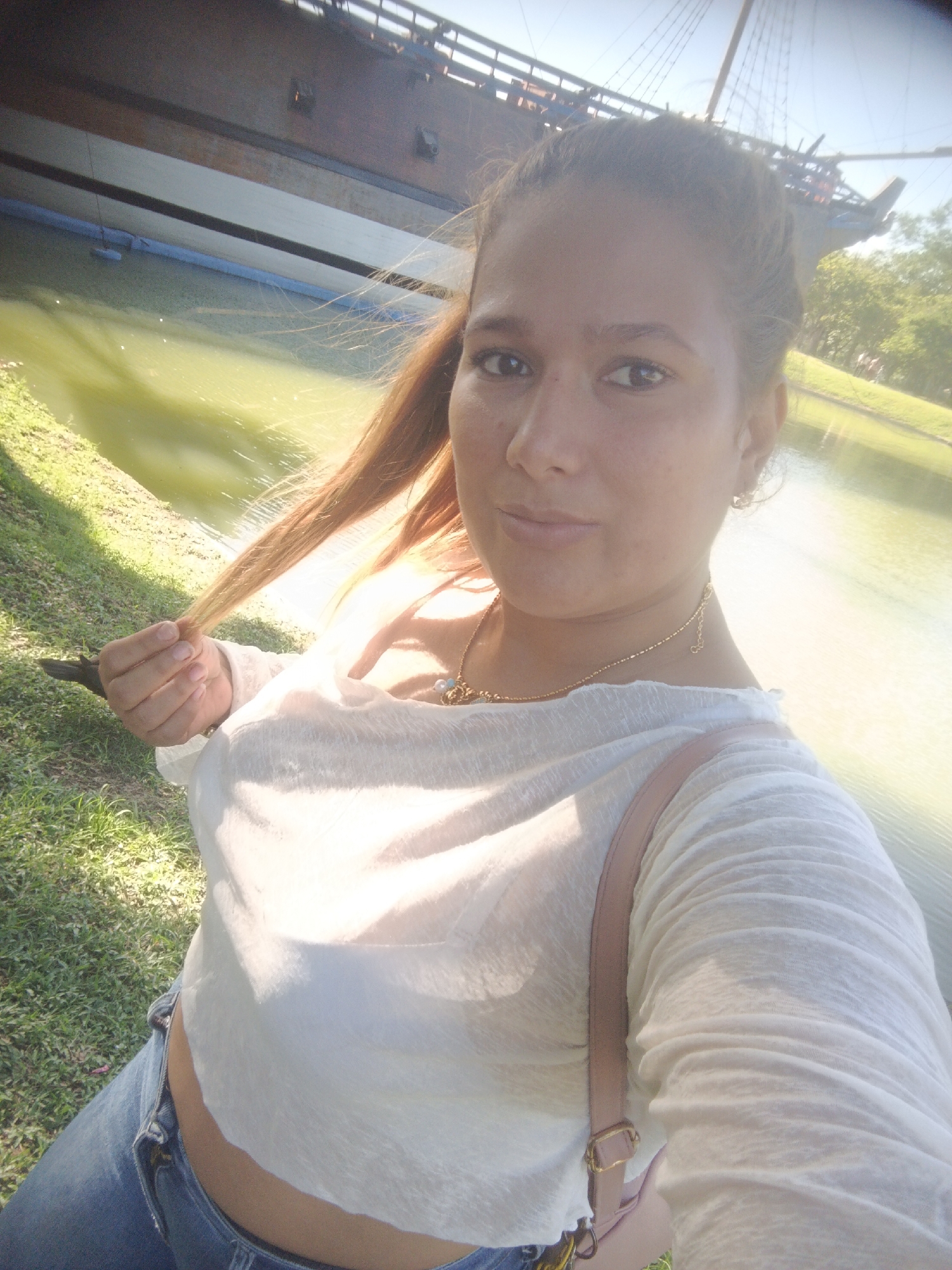 A young woman with long, light brown hair is taking a selfie outdoors. She is wearing a sheer white long-sleeved top over a white bra, and blue jeans. A gold necklace with a pendant is visible. A pink strap from a bag or purse crosses her chest. She is holding a strand of her hair in her left hand. The background shows a body of water, green grass, and trees. In the far background, a large, dark, wooden structure resembling a boat or ship is visible against a bright sky, suggesting an outdoor recreational area, possibly a park or historical site within the city of Caracas, Venezuela, such as the area around the Valencia Lake where such historical replicas might be displayed. The lighting indicates it is daytime, likely sunny.