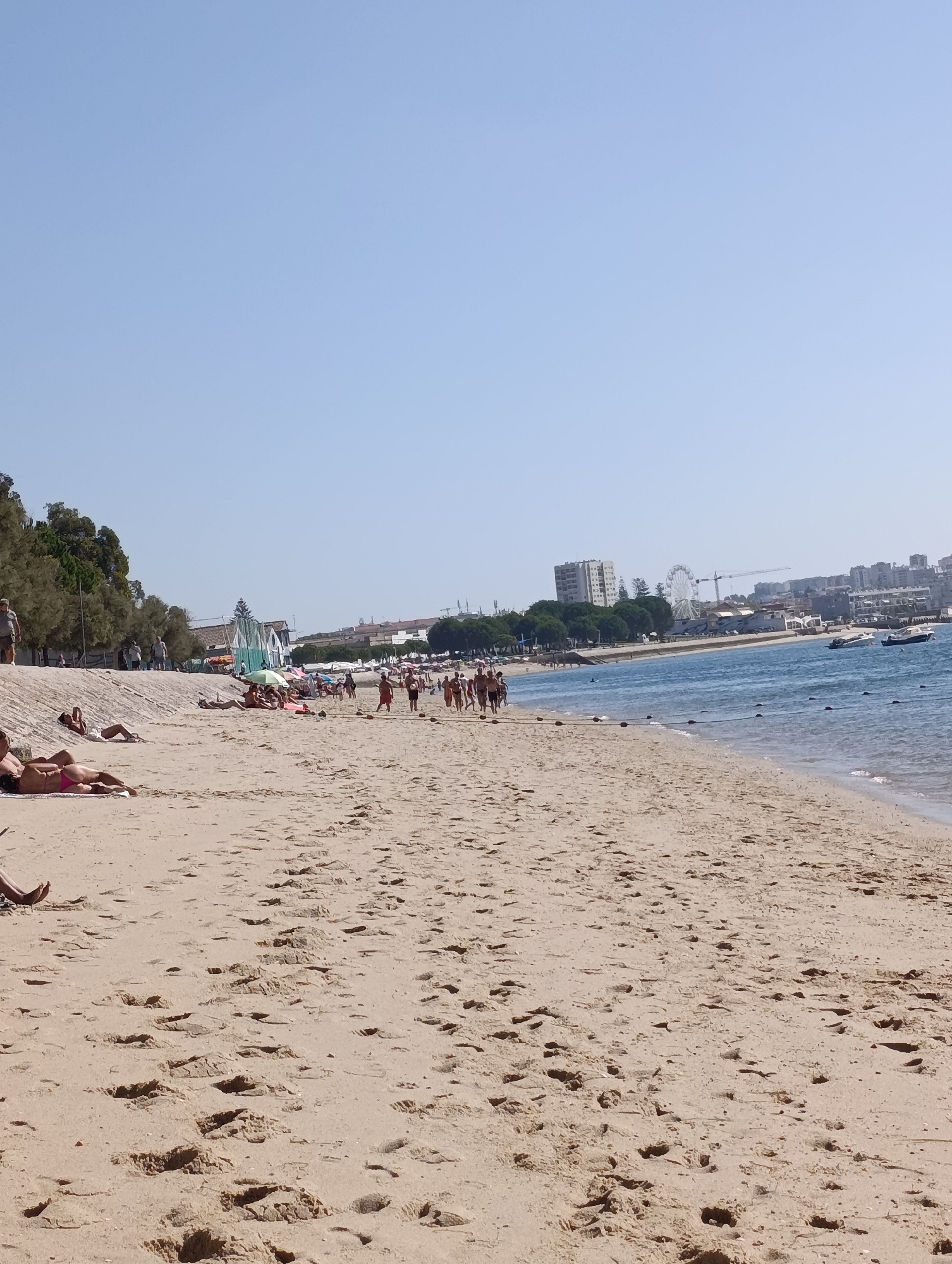 The image shows a crowded beach scene with people sunbathing and walking on the sand. The beach is adjacent to the sea, which has several boats on it. In the background, there are buildings, trees, and a Ferris wheel, suggesting an urban coastal location. The sky is clear and blue. The scene is consistent with the landscape of the Praia da Figueirinha, a popular beach located in the Parque Natural da Arrábida, near the city of Setúbal, Portugal.