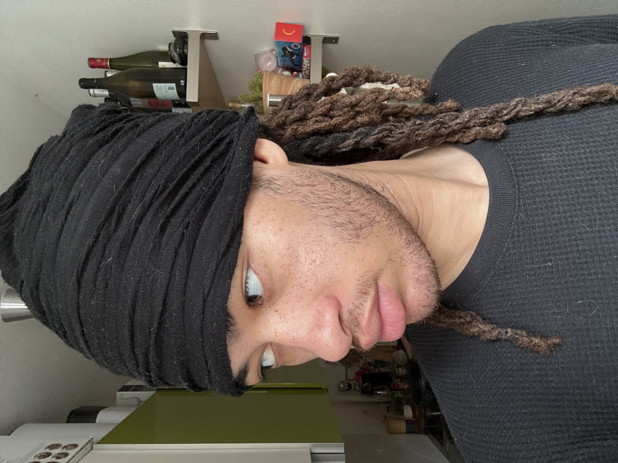 A close-up, upside-down image features a man wearing a black head covering and a black thermal shirt. He has light skin, visible facial hair, and dreadlocks. Behind him are kitchen items, including wine bottles on a rack and miscellaneous containers. The location cannot be determined from the image.
