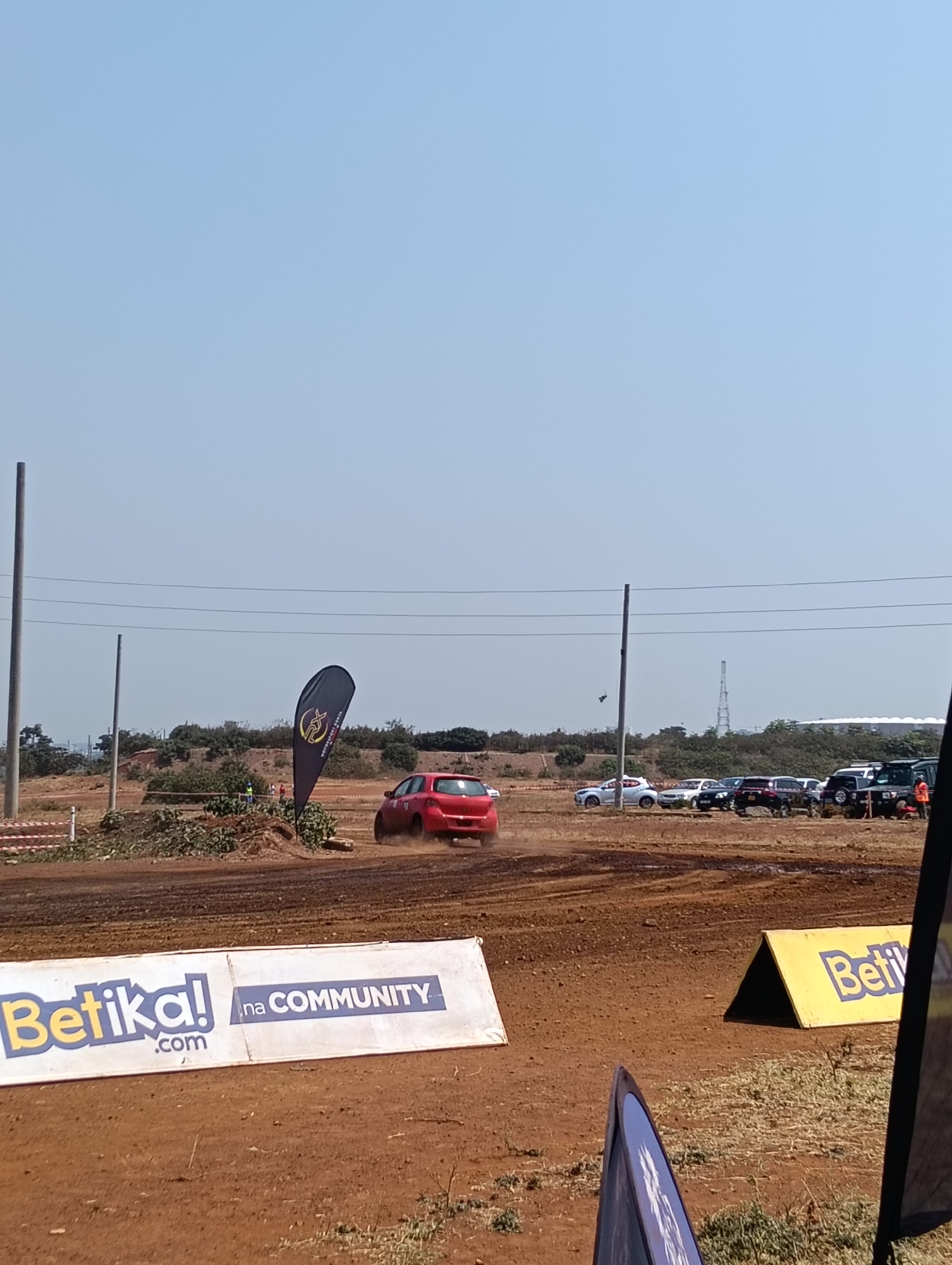 The image depicts a dusty dirt track with several cars participating in what appears to be a rally or motorsport event. In the foreground, a red rally car is seen kicking up dust as it navigates the track. To the left, a white banner with the text "Betika! .com na COMMUNITY" is prominently displayed. Further in the background, a line of parked vehicles is visible, suggesting an audience or staging area. Several tall poles with overhead wires are also present, typical of outdoor event infrastructure. The overall environment is dry and arid, with sparse vegetation and a clear blue sky. The scene likely takes place at a motorsport venue or a designated off-road track within or on the outskirts of Nairobi, Kenya, given the presence of the "Betika!" branding, a Kenyan betting company.
