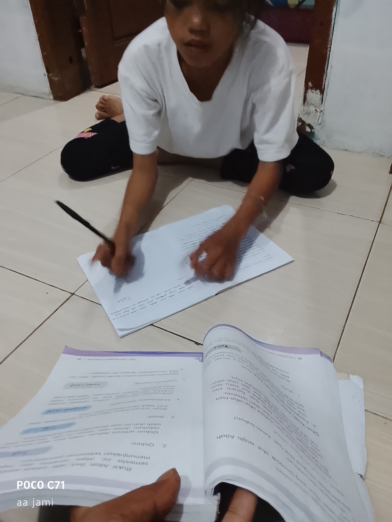 The image shows a child in white clothing sitting on a tiled floor, writing in a notebook with a black pen. The child's legs are bent, and they are wearing black pants. Two open books are placed in front of the child. One book is a textbook with pages displaying text and headings in Indonesian, including "Qaidam" and "Bukti Allah Swt Sifat Wajib Allah". The other book is a notebook where the child is actively writing. The background includes a wooden doorframe and a wall. The lighting suggests an indoor setting. The POCO C71 watermark is present in the lower left corner, along with the text "aa jami".
This scene is likely taking place in a residential area in Tasikmalaya, Indonesia, given the POCO C71 watermark which is a mobile phone brand commonly used in Indonesia, and the presence of Indonesian text in the books. The tiled floor and wooden doorframe are common features in Indonesian homes.