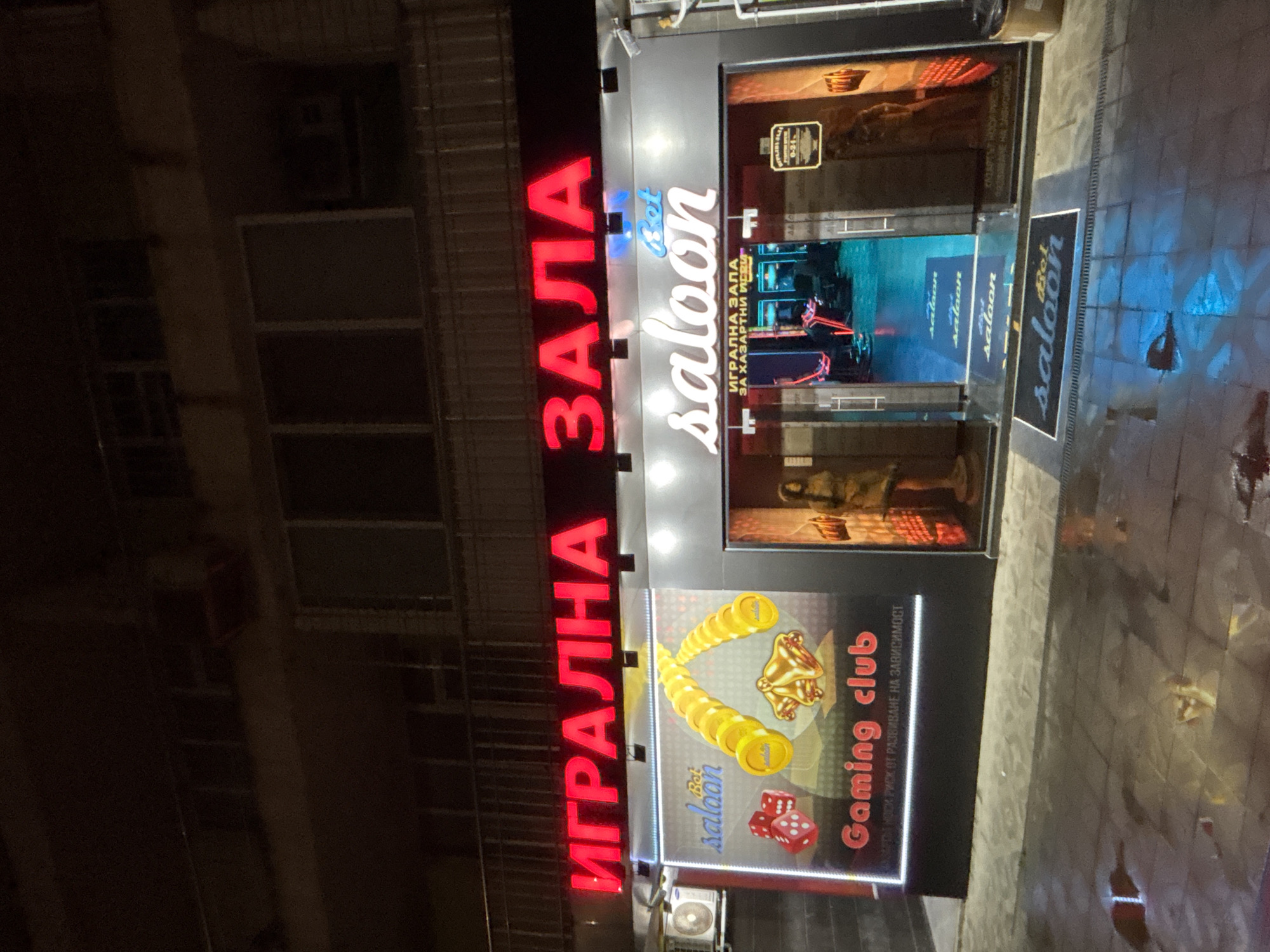 The image shows the exterior of a "Gaming Club" establishment in Pleven, Bulgaria. The sign, written in Cyrillic, says "ИГРАЛНА ЗАЛА" (Gaming Club) with the word "saloon" incorporated into the design. There are images of coins, dice, and a bell, alongside the words "Gaming club" in English. The building itself appears to be older, as evidenced by the aged exterior and architecture, and is located on a city street with wet pavement. There are no people visible in the image. The establishment features a reflective sign on the sidewalk and a light-up display showcasing scenes associated with gambling.