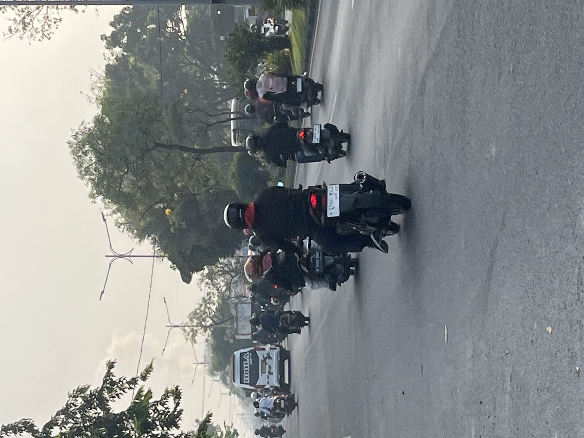 The image depicts a busy street scene in Gelam, Indonesia, likely during daylight hours with overcast skies. A convoy of motorcycles is traveling on a paved road, with some vehicles and pedestrians visible in the background. A white van with the text "IDOLA" is prominently featured in the middle ground. The overall atmosphere suggests a typical urban commute or travel in the region.