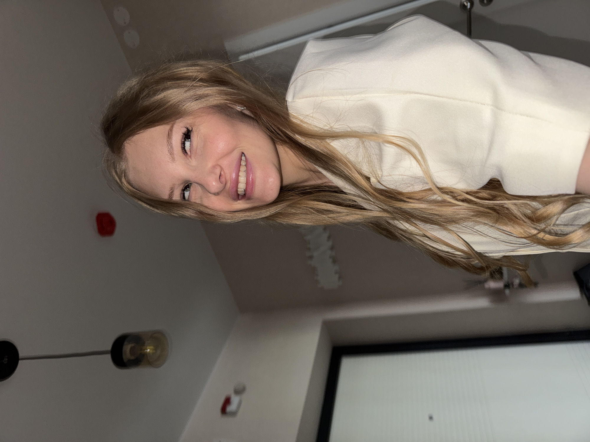 A young woman with blonde hair is shown from the chest up, looking to her left and smiling. She is wearing a cream-colored jacket. The background is an indoor setting with a light-colored wall and a doorway on the right. A light fixture hangs from the ceiling above and to the left. A red object is visible on the ceiling. A closed door with a black frame is to the left of the subject. The scene is likely a residential interior.