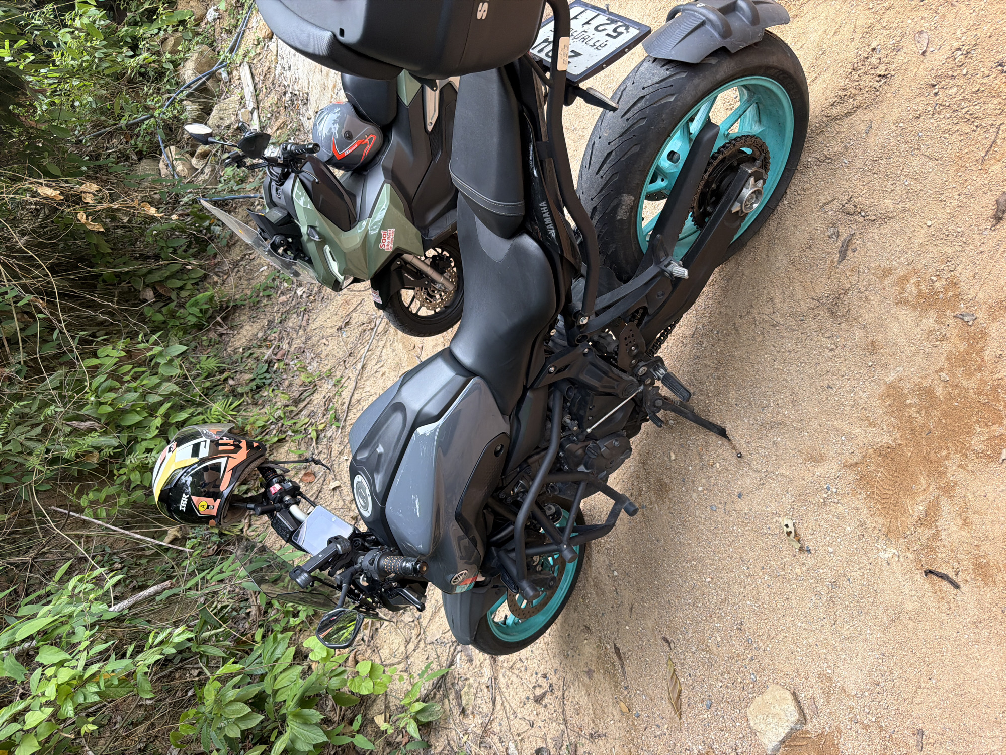 Two motorcycles are parked on a sandy path next to a lush, overgrown embankment. The motorcycle in the foreground is dark gray with bright teal wheels and black crash bars. A helmet with orange, yellow, and white accents is mounted on the handlebars. The motorcycle behind it is a muted green with a black seat and a gray helmet resting on the seat. The environment suggests a natural, possibly rural or mountainous area on the island of Ko Samui, Thailand, given the presence of tropical foliage and the sandy terrain. The scene appears to be a still moment, with no individuals actively engaged in any action.