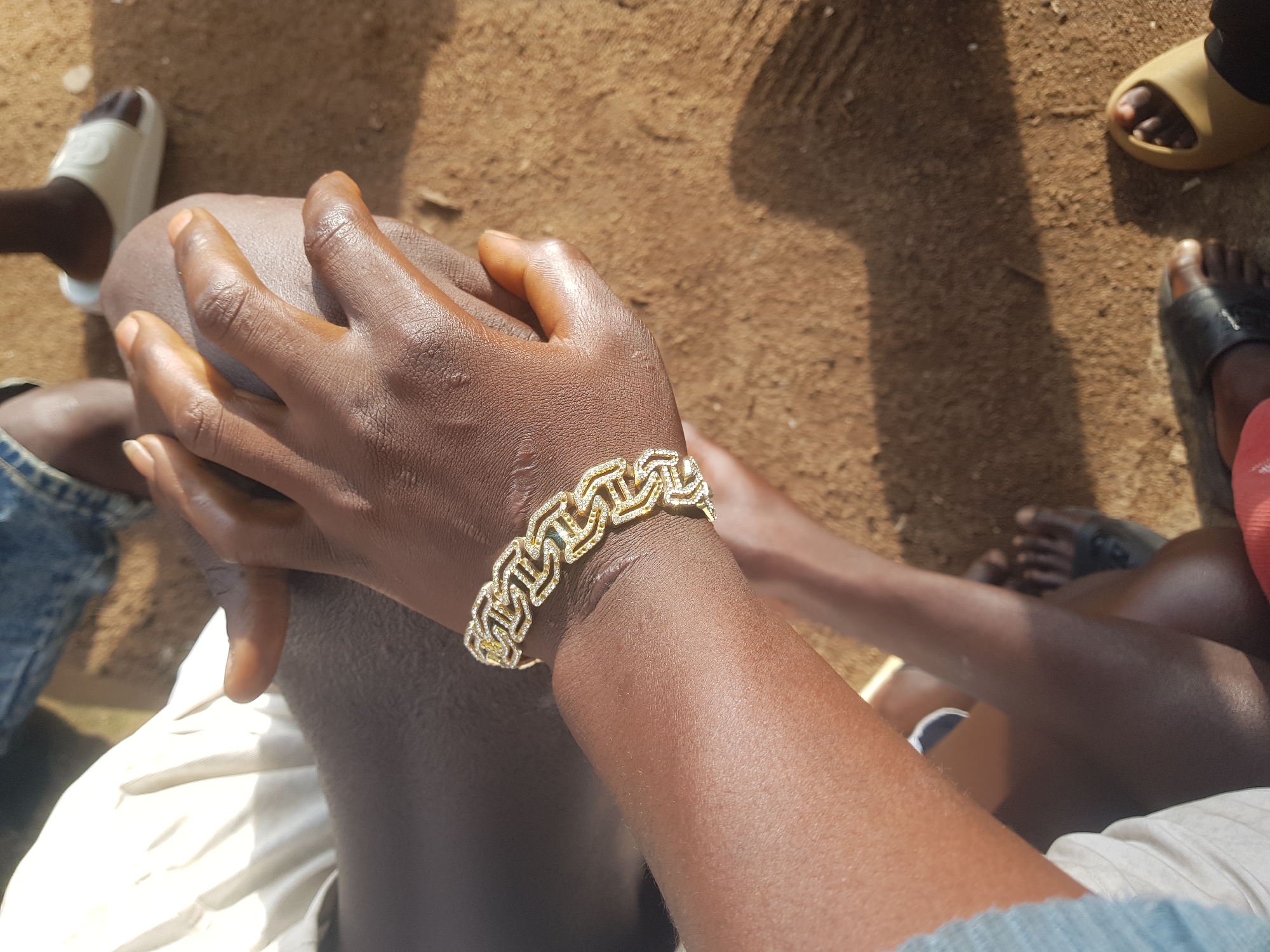 The image shows a close-up view of a person's dark-skinned forearm and hand, adorned with a gold link bracelet studded with small white stones. The person's hand is resting on their knee. The background is a sandy surface, suggesting an outdoor environment. The lighting indicates it is daytime, with distinct shadows cast on the ground. The presence of other individuals' legs and feet, wearing sandals, hints at a gathering or social setting. Based on the context of Jos, Nigeria, this scene might depict a casual moment in a public space or private gathering within the city.