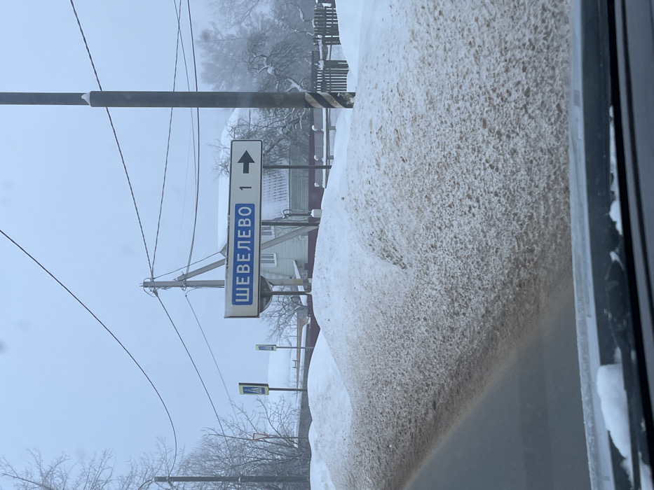 The image depicts a winter street scene in Belavino, Russia, viewed from inside a vehicle. A significant dirty snowdrift borders the right side of a cleared road, extending upwards from the lower left frame. A rectangular road sign, displaying "ШЕВЕЛЕВО 1" (Shevelevo 1) in blue Cyrillic text with a right-pointing arrow on a white background, is prominently positioned above the snowdrift. Multiple utility poles with overhead wires traverse the scene. In the midground, a light green house with a dark roof is partially visible behind the sign and infrastructure. Further down the road to the left, two yellow pedestrian crossing signs are discernible. A dark wooden fence is visible behind the snowdrift and in front of the trees. The background features bare deciduous trees, some lightly coated in snow or frost, receding into a light atmospheric haze under an overcast, light grey sky. The vehicle's dashboard and the lower edge of the windshield, with minor snow accumulation on the wipers, are visible in the foreground. No individuals are present.