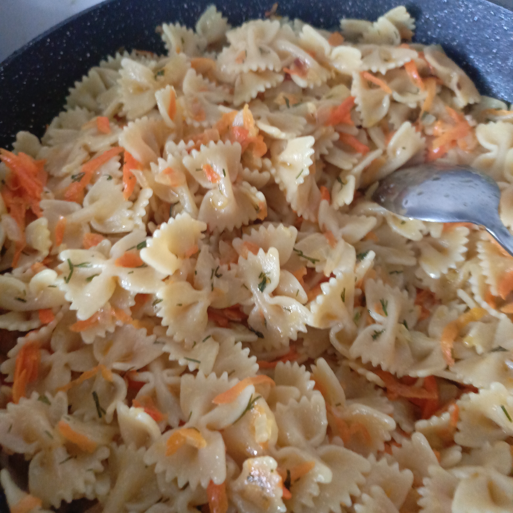 The image presents a close-up view of a cooked pasta dish contained within a dark-colored pan, possibly a skillet or wok. The primary component of the dish is farfalle pasta, characterized by its distinctive bow-tie shape and light beige or yellowish hue. Interspersed generously among the pasta are vibrant orange pieces, which appear to be shredded or finely diced carrots. Small, dark green specks, resembling chopped herbs such as dill or parsley, are also visible throughout the dish.
The pasta and vegetables are coated in a light, glossy sauce or oil, suggesting they have been sautéed or tossed together. A metallic silver spoon is partially visible on the right side of the frame, resting within the pasta. The pan's interior edge, visible at the top and left, is dark grey or black with a speckled non-stick texture. The scene depicts a meal being prepared, stirred, or served. No people are visible, and there is no discernible text within the image. The setting is likely an indoor environment, such as a kitchen.