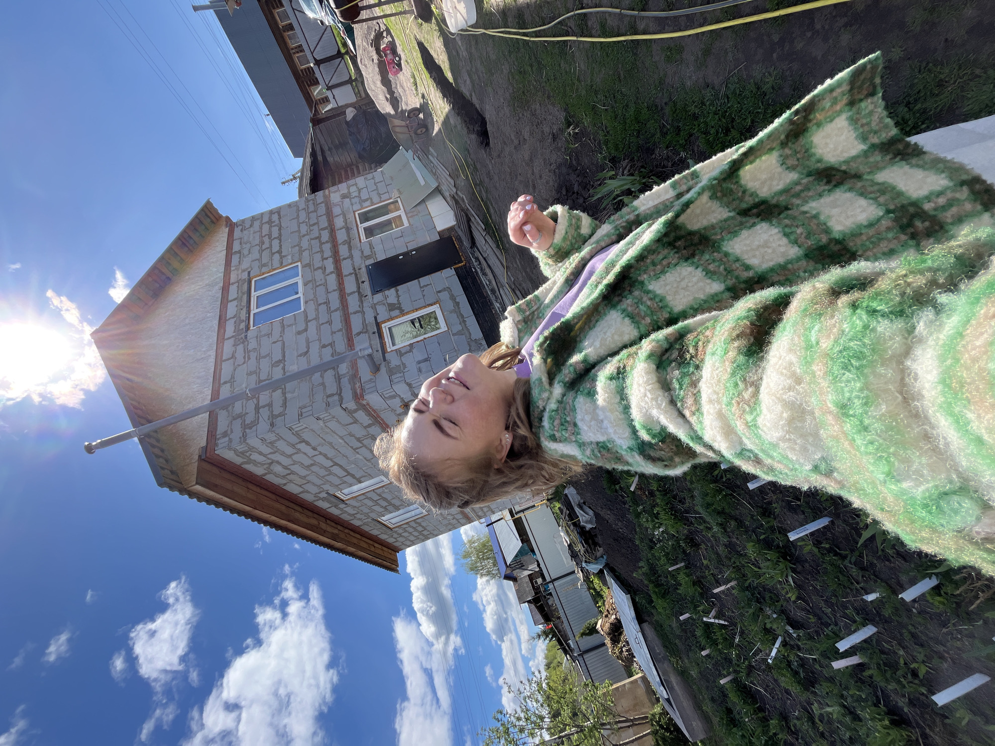 A woman wearing a green and white plaid jacket is posing for a selfie in front of a partially constructed brick building. The location is identified as Ольховка, Russia. The building appears to be residential, with windows installed and a door visible. Behind the building is a yard with a garden or planted area. The sky is blue with scattered white clouds. A yellow hose is lying on the ground.