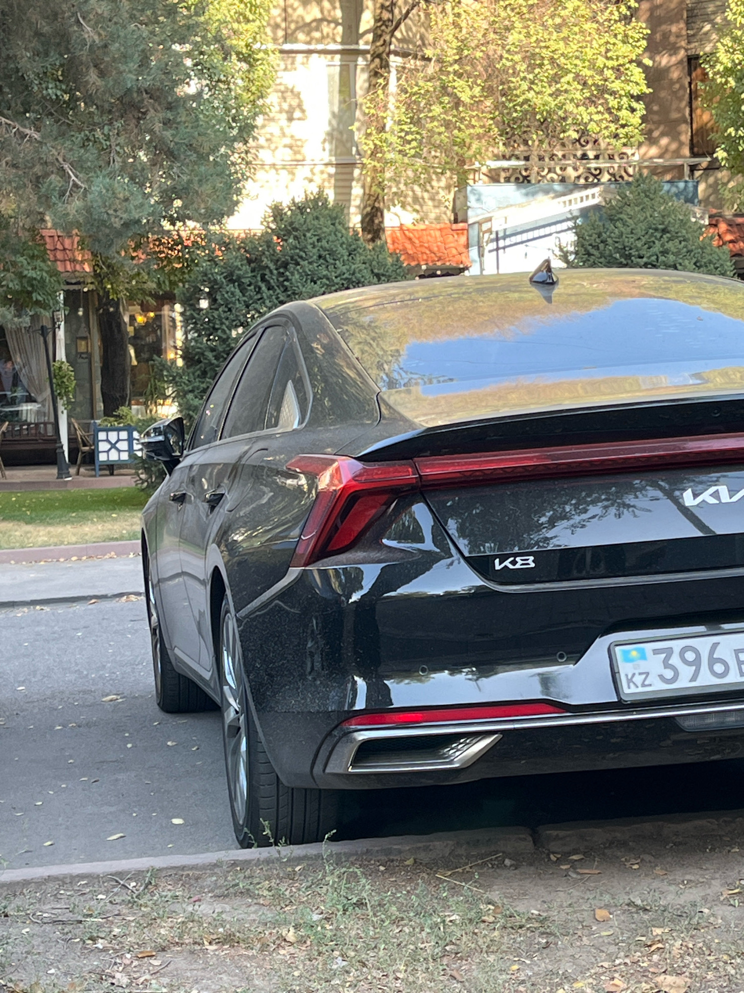 The rear left quarter of a black Kia K8 sedan is parked on a street in Almaty, Kazakhstan. The license plate reads "KZ 396 B". The car is positioned at an angle, with its left side closer to the camera. In the background, there are trees, a building with red-tiled roofs, and a fence. The street is paved and appears to be lined with trees and grass. The overall impression is of a daytime urban scene.