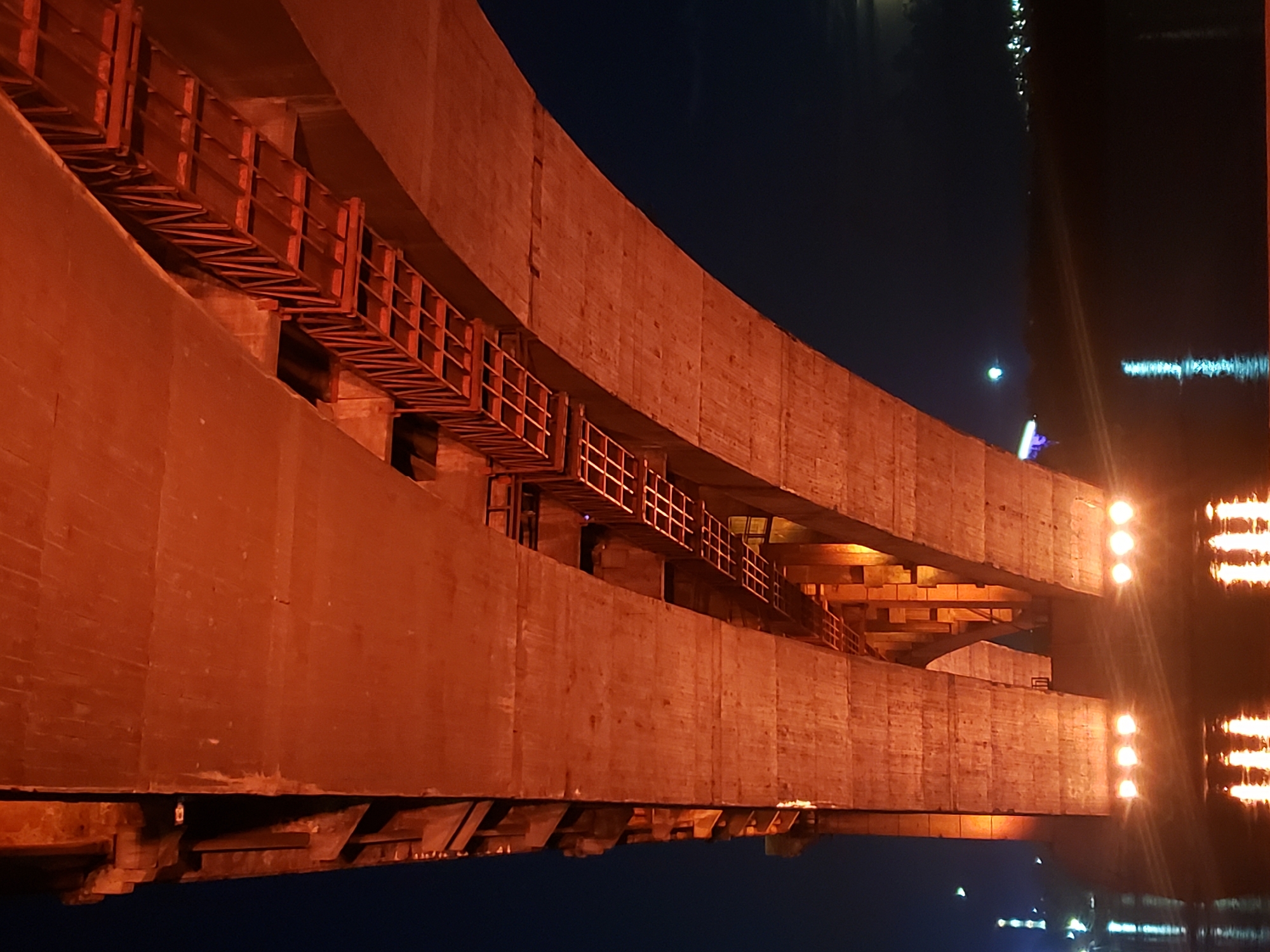 The image shows a close-up of a large, curved concrete structure at night, likely a bridge or overpass. The concrete is illuminated by warm, orange lights, creating a dramatic and somewhat ominous atmosphere. In the foreground, a section of the structure reveals a metal railing and walkway, suggesting it is a pedestrian or maintenance access. This walkway appears to be suspended beneath the main concrete structure, creating multiple levels within the architecture.
The background is a deep, dark blue, indicating nighttime. Reflections of lights are visible on a dark surface to the right of the frame, possibly water, hinting that the structure is elevated above a body of water or a wet surface. A few bright, white lights are also visible on the concrete structure itself, some in distinct rows, adding to the artificial illumination of the scene. The overall impression is one of scale and industrial architecture against the vastness of the night sky. There is no visible text in the image. The scene evokes a sense of solitude and quiet grandeur.