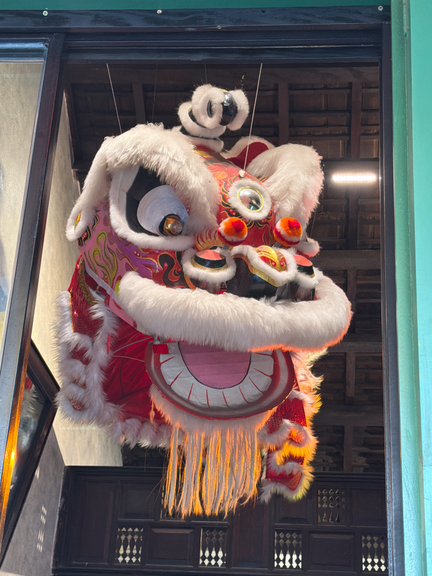 A decorative lion head, typically used in lion dances during festivals, is suspended in the air inside what appears to be an entryway or open doorway. The lion head is elaborately designed with red, pink, and white fur, and gold and black accents. It has large, reflective eyes and a wide, open mouth with pink padding. Golden tassels hang from the bottom of the head. The background reveals a wooden ceiling structure and a bright light, possibly a fluorescent tube. Below the lion head, a dark wooden lattice-like structure is visible. The scene is framed by dark window or door frames, with a sliver of a light-colored wall on the left and a green painted wall on the right. The overall impression is one of festive decoration, potentially for Lunar New Year or a similar celebration. Based on the architectural style of the interior, particularly the wooden ceiling and the lattice work, this scene could be located in an older building or a place designed to evoke traditional Vietnamese aesthetics. Given the detail of the lion dance costume, it is highly likely this is in Vietnam, and Nha Trang is a major city in Vietnam.