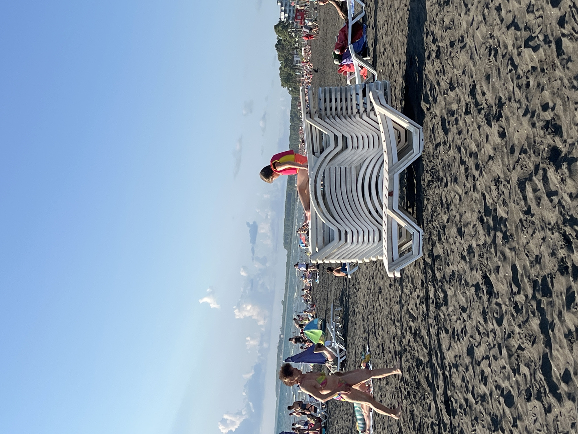 This image depicts a beach scene in Ureki, Georgia, characterized by dark sand and a clear blue sky. In the foreground, a stack of white plastic lounge chairs is prominently displayed. To the left of the chairs, a woman in a colorful bikini walks across the sand. Further back, other beachgoers are visible, some lounging on chairs or towels, while others are in the water. The middle ground shows a line of trees and buildings along the coastline, suggesting a developed resort area. A person wearing a bright pink and yellow life vest is seated on top of the stacked lounge chairs. The overall atmosphere is that of a sunny day at a crowded beach.