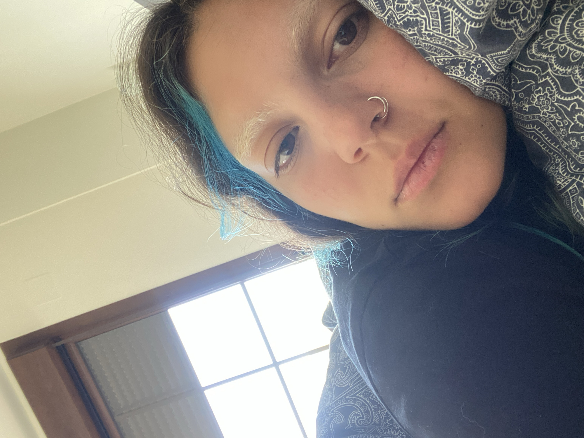 A close-up, indoor shot features a person with fair skin, brown eyes, and a nose ring. The person has dyed blue streaks in their dark hair and bleached eyebrows. They are partially obscured by a gray patterned fabric, likely clothing or a blanket, and wearing a black garment. A window with a grid pattern is visible in the background, allowing bright sunlight to stream in. The setting does not provide sufficient clues to pinpoint a specific location within Villaviciosa, Spain.