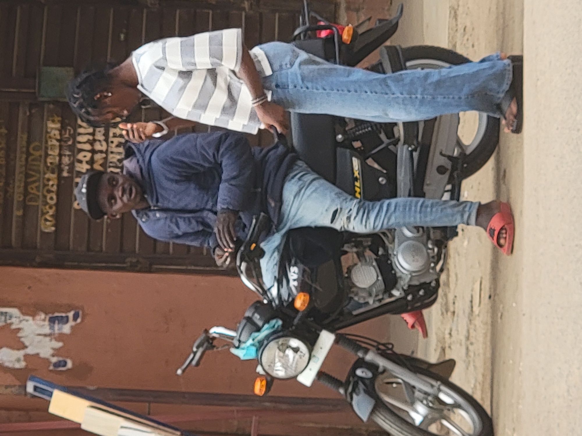 Two young men are interacting with a TVS motorcycle in an urban environment, possibly Maiburiji, Nigeria. One man, wearing a striped shirt and jeans, stands next to the motorcycle, looking at a mobile phone. The other, wearing a blue jacket, jeans, and a baseball cap, is sitting on the motorcycle. The motorcycle is parked on a paved surface next to a building.
