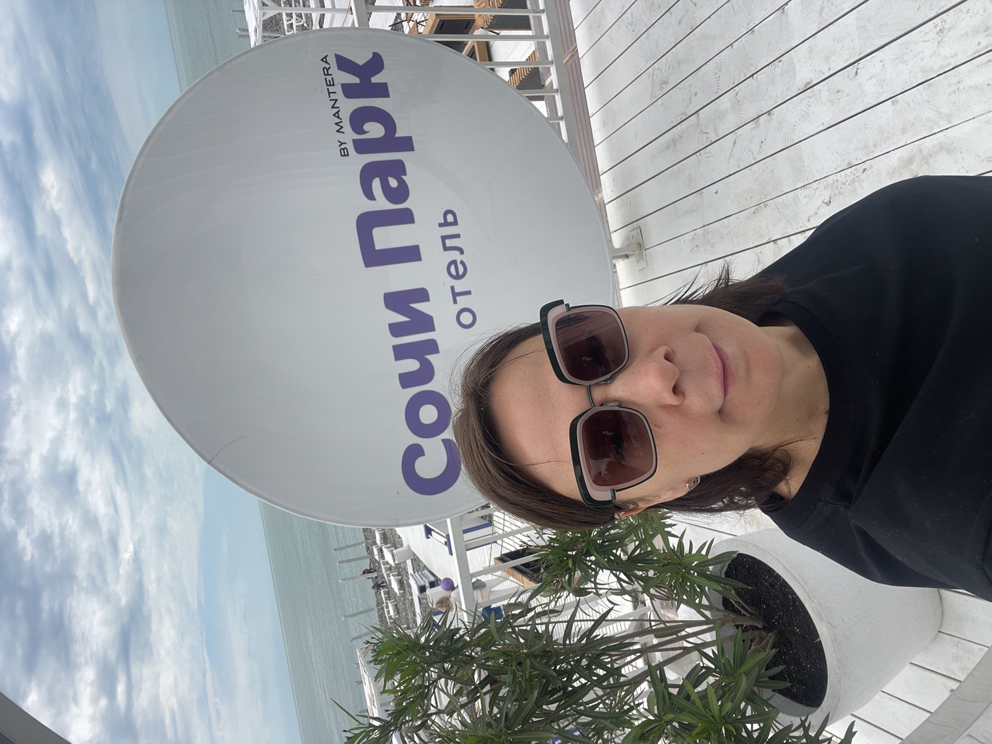 A person wearing sunglasses and a black shirt is standing next to a large, round sign displaying the words "Сочи Парк Отель" and "BY MANTERA". The sign is likely affiliated with the Sochi Park Hotel in Sochi, Russia. The person is positioned near a potted plant with green foliage. In the background, a body of water, possibly the sea, is visible, along with a white structure that may be part of the hotel or a surrounding facility. The image appears to be a selfie taken from a high vantage point.