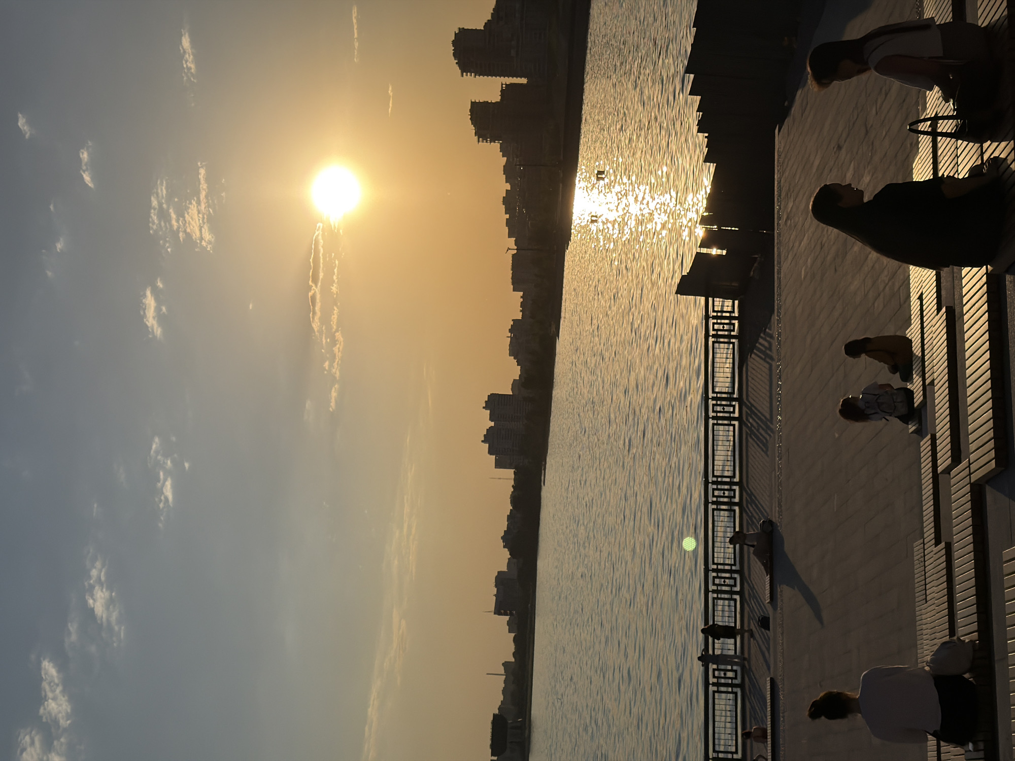 The image shows a sunset scene in Kazan, Russia. The sun is setting over the city skyline, reflecting off the water. A walkway runs along the right side of the water with people sitting, standing, and walking. A metal railing separates the walkway from the water.