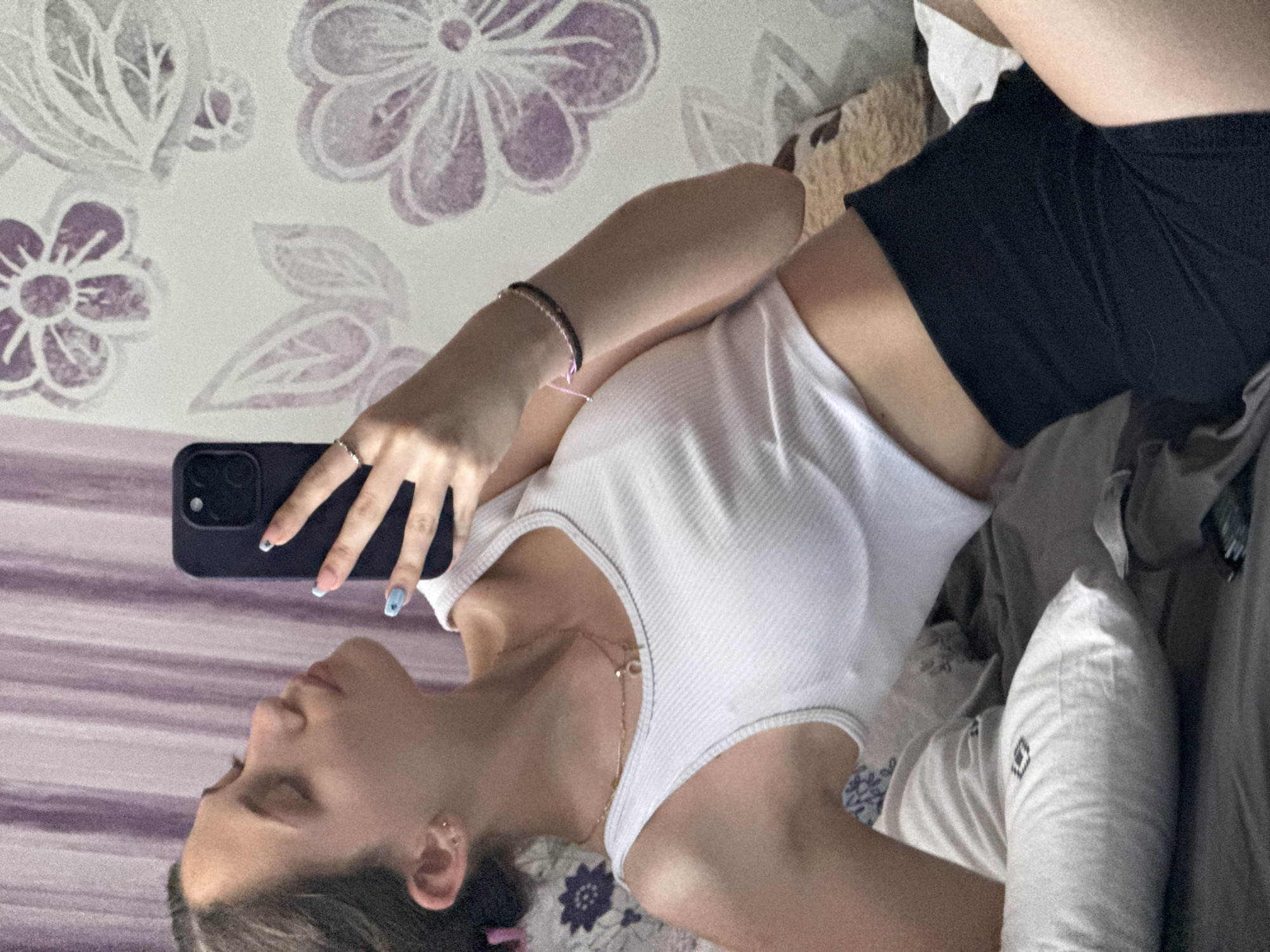 A young woman is taking a mirror selfie in what appears to be a bedroom. She is wearing a white ribbed tank top and black shorts. She has on a necklace and a bracelet. Her nails are painted blue with white designs. The background shows a wall with a floral pattern and a purple curtain. The image is taken from a high angle, showing her from the shoulders up and her torso. The overall lighting suggests it is daytime. The location is not specified in the image, and it is not possible to determine if this is in Sofia, Bulgaria.