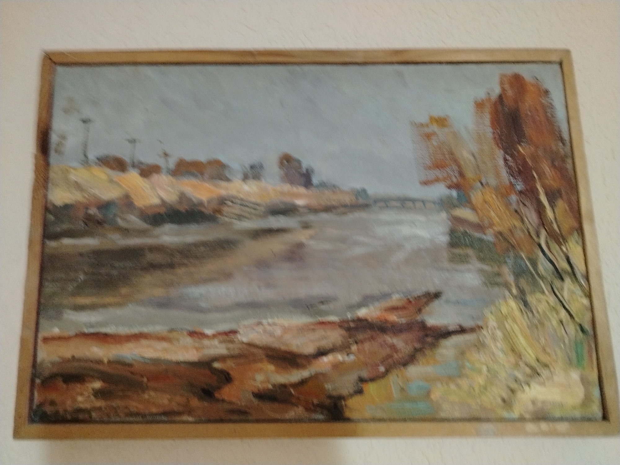 The image is a framed oil painting depicting a landscape scene. In the foreground, there is a body of water, possibly a river or canal, with a rocky or muddy bank on the left side. The water reflects the sky and the surrounding landscape.
The middle ground shows a flat area with sparse vegetation and what appear to be structures or landforms. There are hints of buildings or industrial elements in the distance, suggested by vertical lines that could represent smokestacks or utility poles. The landscape appears somewhat barren, with patches of yellow and brown earth.
The background is dominated by a hazy, overcast sky, rendered in shades of gray and pale blue. On the right side of the painting, there are several trees or bushes with autumnal foliage, painted in vibrant shades of orange, brown, and yellow. A distant bridge or causeway is visible spanning the water.
The overall style of the painting is impressionistic, with visible brushstrokes and a focus on capturing the atmosphere of the scene rather than precise detail. The painting is presented in a simple wooden frame. There is no indication in the image or its context that this scene is specifically located in the city of Nukus, Uzbekistan.