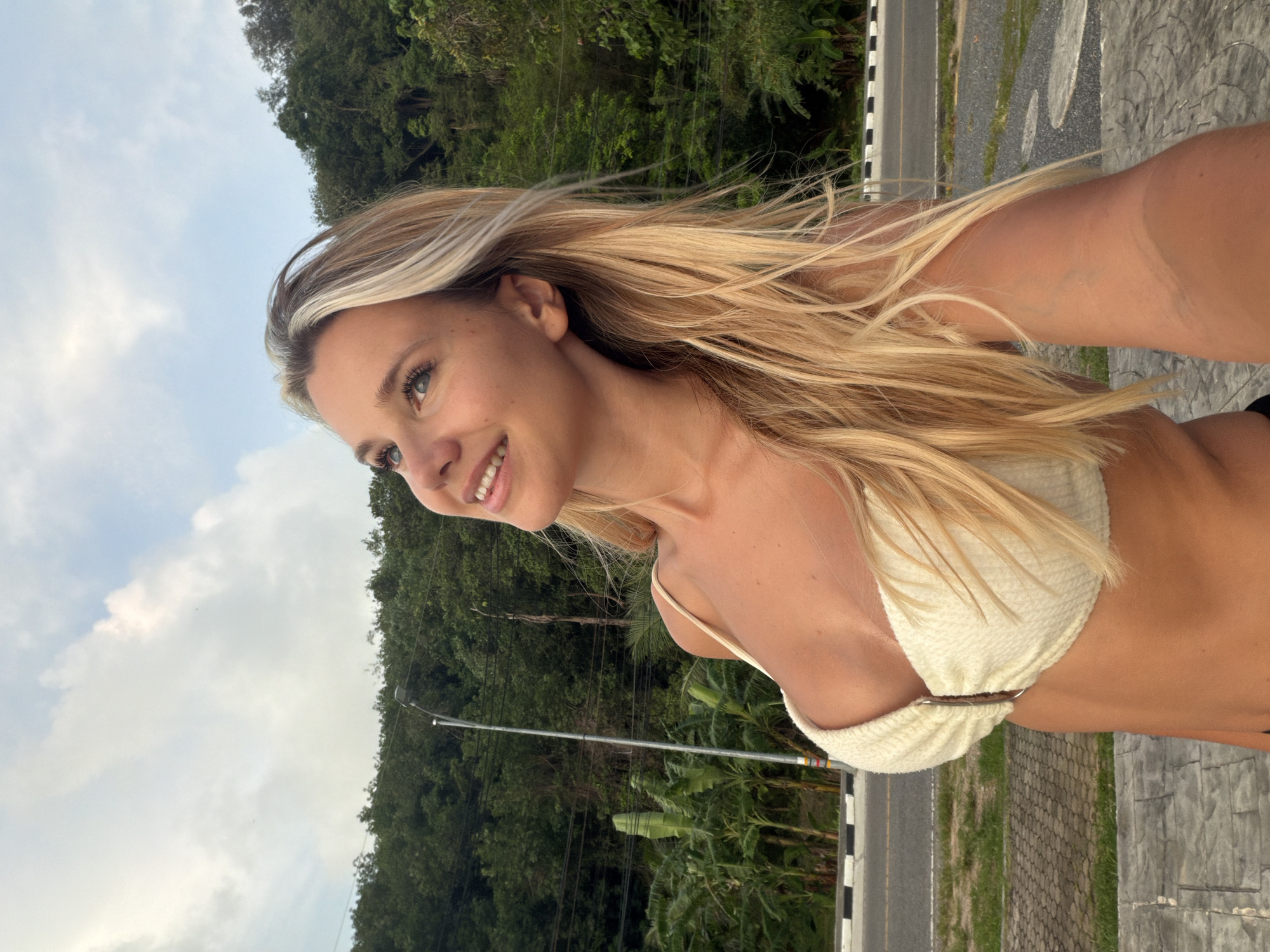 A young woman with long blonde hair and blue eyes, wearing a cream-colored bikini top, poses for a selfie outdoors. She is looking upwards and to her left with a slight smile. Behind her, a lush green hillside covered in trees rises above a road with a white and black striped curb. Banana trees are visible on the left side of the image. The ground in the foreground is paved with grey hexagonal tiles. The scene appears to be in a tropical or subtropical location, possibly Wichit, Thailand, given the vegetation and road infrastructure. The sky is partially cloudy.