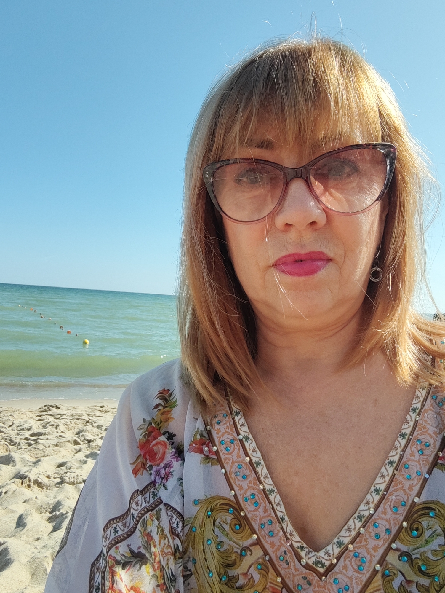 A woman with blonde hair and bangs, wearing oversized cat-eye glasses and a floral-patterned sheer dress, poses for a selfie on a sandy beach. The woman's lips are painted a bright pink, and she wears a circular earring in her right ear. Behind her, the turquoise sea meets a clear blue sky, with a rope line marked by buoys in the water. The foreground shows the sandy beach with shadows cast by unseen objects. The image appears to be taken on a sunny day. The location is identified as the city of Zelenogradsk, Russia.