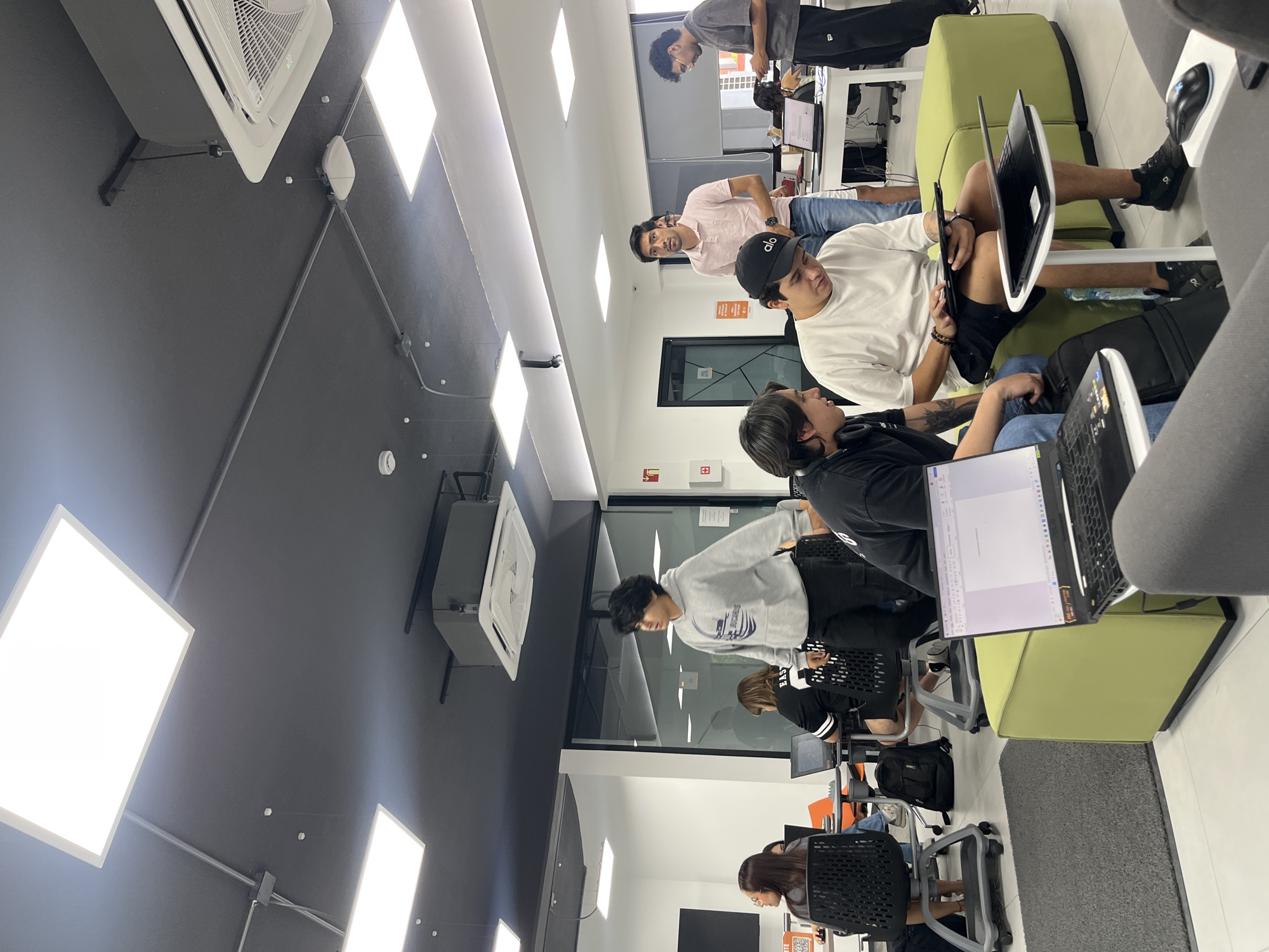 The image depicts an indoor office or co-working space environment in San Andrés Cholula, Mexico. Several individuals are present, engaged in work-related activities. Some are seated on lime-green couches while using laptops and tablets, while others are sitting at desks using computers. One man wears a black baseball cap and a white t-shirt, looking down at a tablet. Another man near him wears a black t-shirt and headphones, focusing on his laptop screen. Further in the background, additional people are working at desks. The room has a black ceiling with rectangular LED panel lights and air conditioning units. The overall atmosphere suggests a casual but productive workspace.