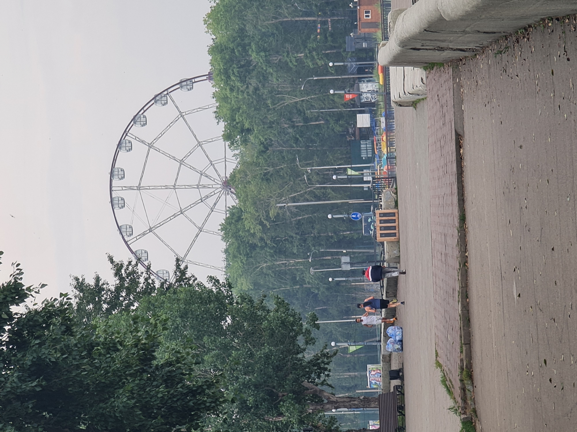 In an outdoor park setting, a large Ferris wheel with a white spoke structure and transparent cabins is visible in the background against a hazy, overcast sky. In the mid-ground, a dense line of green trees forms a backdrop. In the foreground, a paved walkway curves to the right, bordered by a rough, concrete wall. Three individuals are jogging on the walkway. The person on the left is wearing a white tank top and shorts, the person in the middle is wearing a dark blue tank top and shorts, and the person on the right is wearing a red jacket, dark pants, and a hat. To the left of the joggers, a dark park bench, some garbage bags, and a colorful mural are present. Further to the right, several small, colorful paddle boats are lined up, and beyond them are some small wooden structures and lights. The ground is covered in a mix of paved surfaces, cobblestones, and scattered leaves.