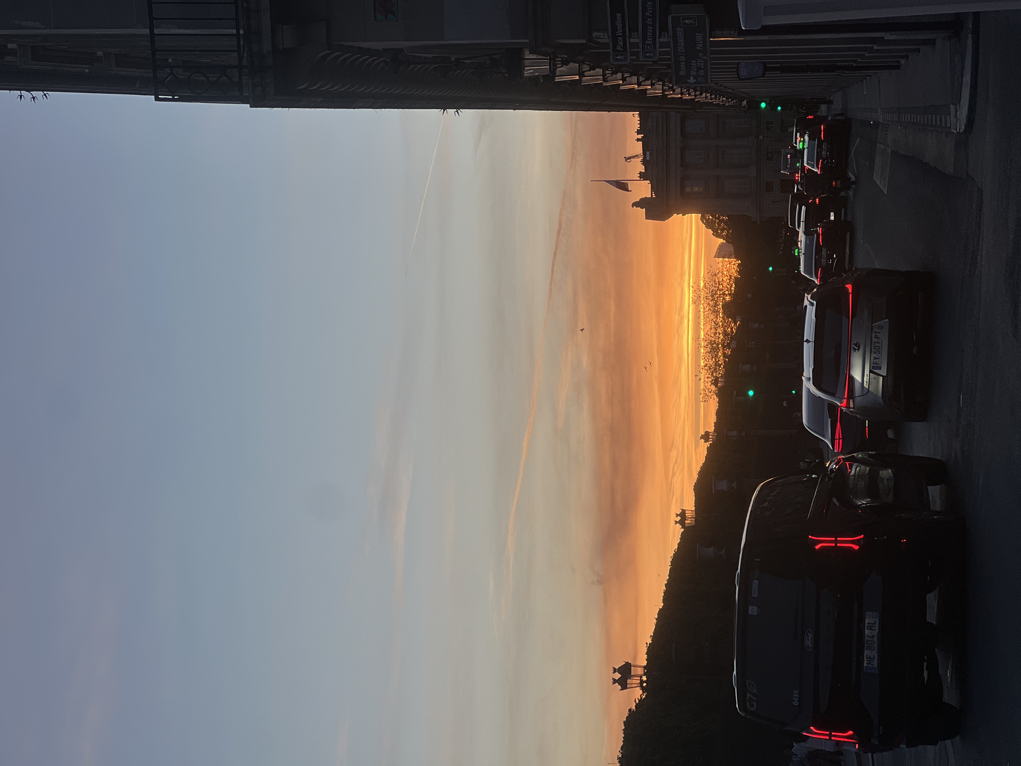 The image presents a vibrant urban landscape in Paris, France, captured at sunset. The photograph, ideally viewed rotated 90 degrees counter-clockwise to correctly orient the street at the bottom, depicts a bustling city street overlooking a body of water, likely the River Seine.
In the foreground, a queue of dark-colored vehicles, including a black Ford van bearing "G70" and "0439" along with the license plate "HE-804-RL", are visible on the road. Another car shows the license plate "FY-507-PT". Red brake lights illuminate the immediate foreground, while green traffic lights are discernible further along the street. Informative street signs point to iconic Parisian locations such as "Place Vendome," "Bureau de Poste," "Musée de l'ORANGERIE," and "Musée du JEU DE PAUME," with an additional indicator of "320 m" to "PAUME."
The midground is dominated by the spectacular golden glow of the setting sun, which reflects intensely off the water's surface, creating a sparkling, almost liquid gold effect. The opposite bank appears in dark silhouette, revealing a dense line of trees, distant architectural forms, and a flagpole flying a flag, likely the French national flag.
The sky, filling the background and top portion of the frame, transitions beautifully from a pale blue at its zenith to a warm blend of orange, pink, and yellow closer to the horizon. A distinct white contrail from an aircraft slices across the upper sky, accompanied by faint wispy clouds. The top edge of the image is framed by the dark silhouette of a building with visible balconies. The overall scene captures the serene beauty of a Parisian sunset amidst the city's daily rhythm.