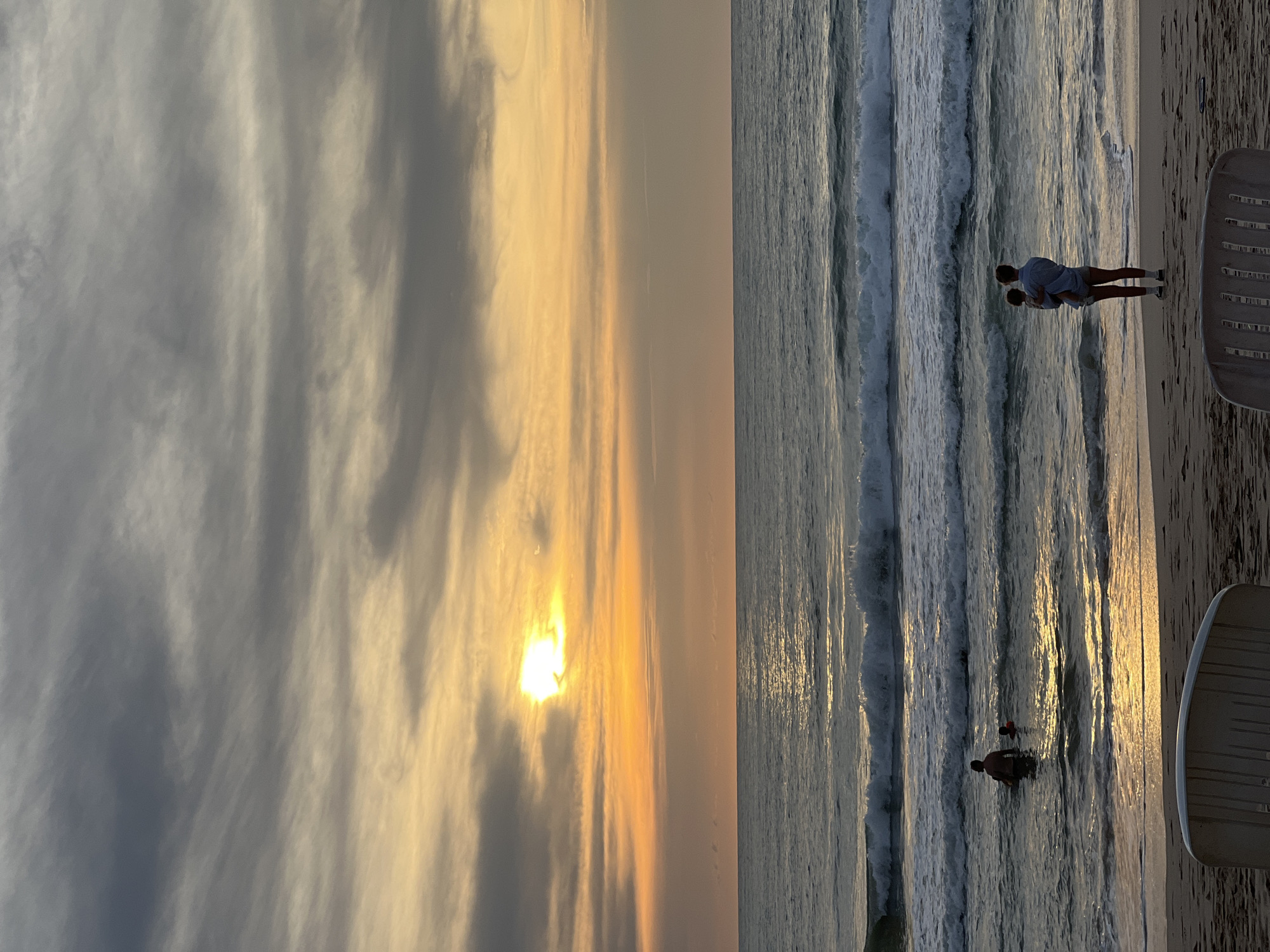 A serene sunset unfolds over the ocean in Choeng Thale, Thailand. The sky is a canvas of warm yellows and oranges, streaked with dramatic grey clouds, as the sun, partially obscured by clouds, casts a radiant glow on the water's surface. Gentle waves roll towards a sandy beach, where two individuals stand near the water's edge. One adult, holding a child, gazes out at the vast expanse of the sea. Further out in the water, another person is visible, wading in the gentle surf. The foreground of the image shows the sandy beach with two chairs.