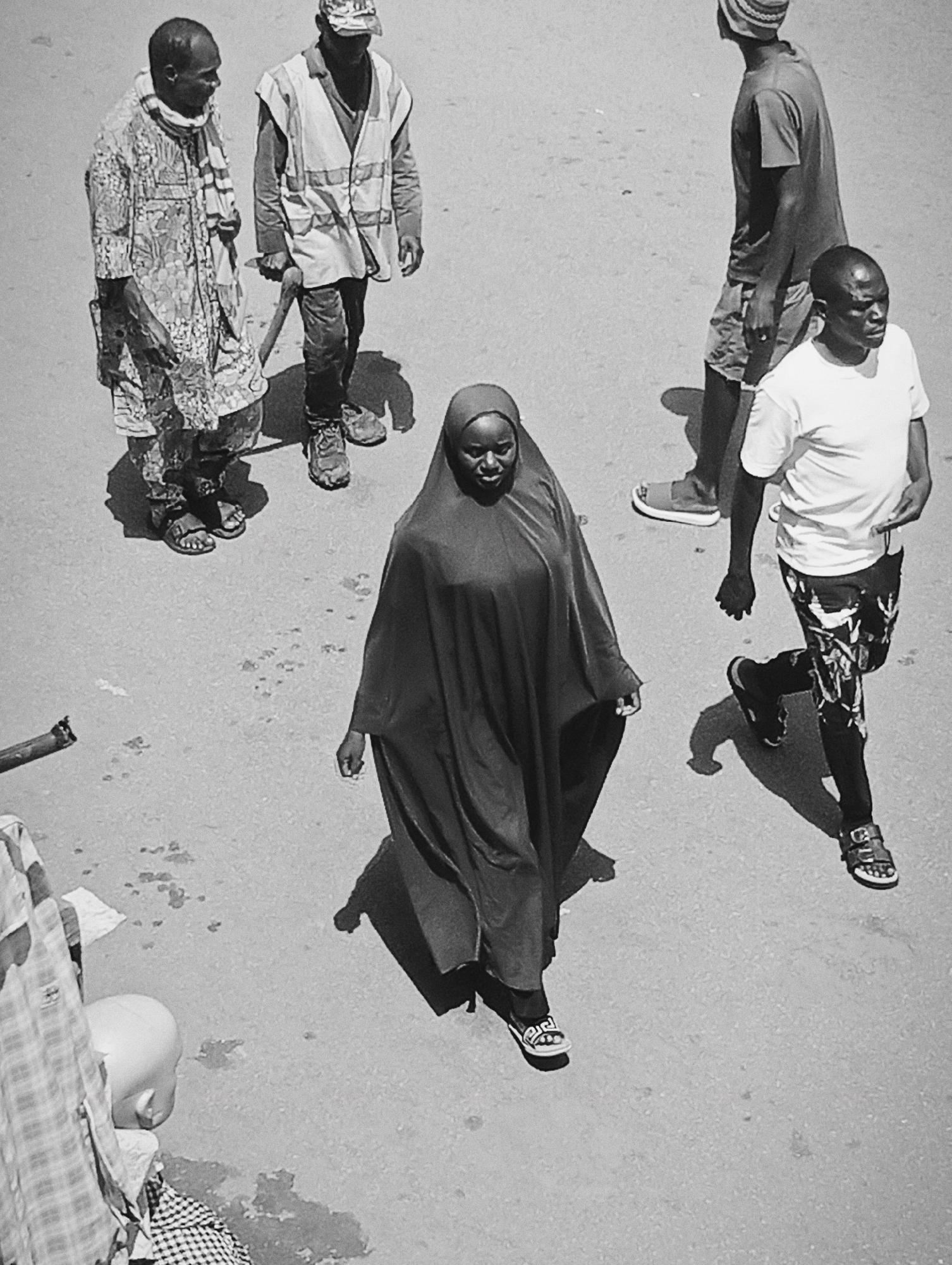 In Bukuru, Nigeria, a group of individuals walk across a dusty, paved surface. In the foreground, a person wearing a dark head covering and a flowing, long garment walks towards the right of the frame. To their left, another person in a patterned robe and a scarf also walks towards the right. Behind them and to the right, a person in a white t-shirt and patterned shorts walks towards the viewer. Further back and to the left, two men are walking away from the viewer. One wears a reflective vest over a long-sleeved shirt, holding a tool that resembles a pickaxe. The other man, further left, wears a patterned robe and a scarf. The environment appears to be an outdoor urban setting, possibly a street or marketplace, with the ground showing signs of wear and dirt.
