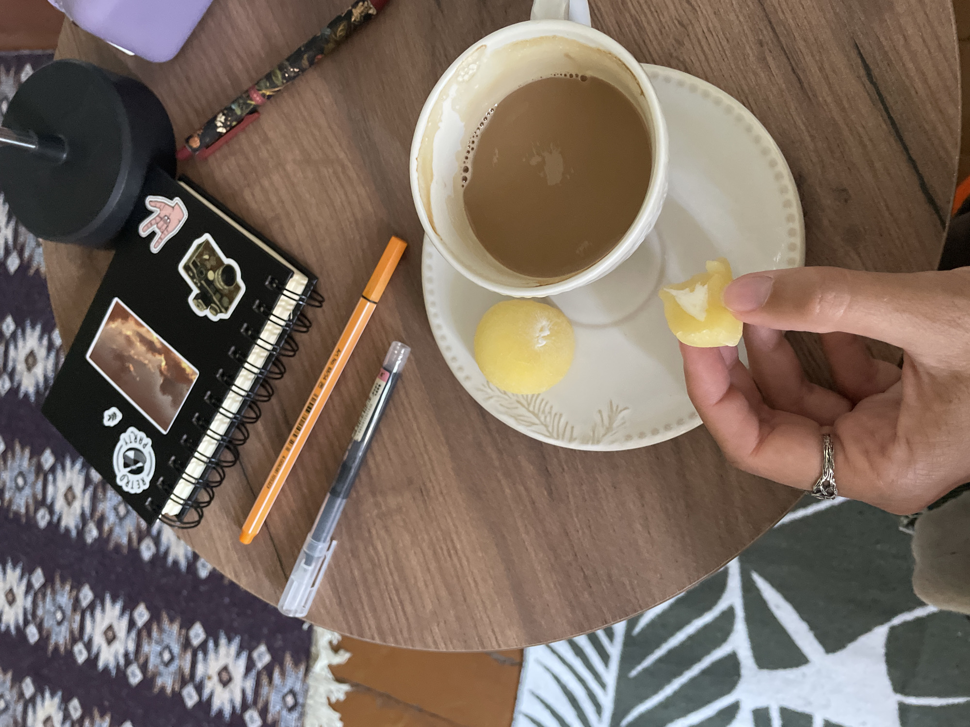 A close-up, overhead shot shows a hand holding a half-eaten yellow mochi over a white saucer. A white teacup filled with coffee sits on the saucer. The coffee has a layer of foam and appears to be mixed with milk. To the left of the cup, on the saucer, is an intact yellow mochi. A spiral-bound black notebook with several stickers is visible in the upper background. Two pens, one orange and one black, lie next to the notebook. A dark, patterned rug is in the background. The scene is set on a round, wooden table. The lighting suggests an indoor setting, possibly a home or cafe.