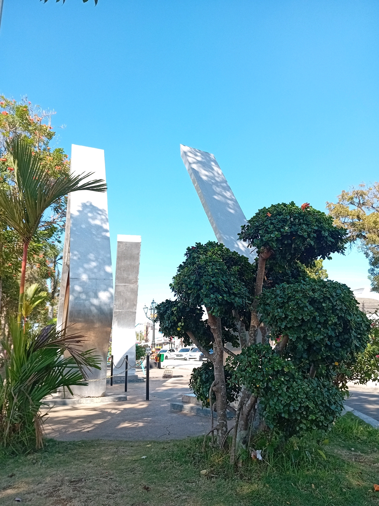The image depicts a section of a public park in Purworejo, Indonesia, featuring several abstract, tall, light-colored monuments that appear to be made of concrete or metal. The monuments are of varying heights and angles, with one leaning dramatically. In the foreground, a lush, manicured green tree with small red flowers is prominently positioned. To the left, a palm tree adds a tropical element to the scene. Beyond the monuments, a street is visible with cars and what appear to be streetlights. The sky is a clear, bright blue, indicating sunny weather. The ground in the park is a mix of grass and paved walkways. There are no people clearly visible in the immediate foreground or midground.