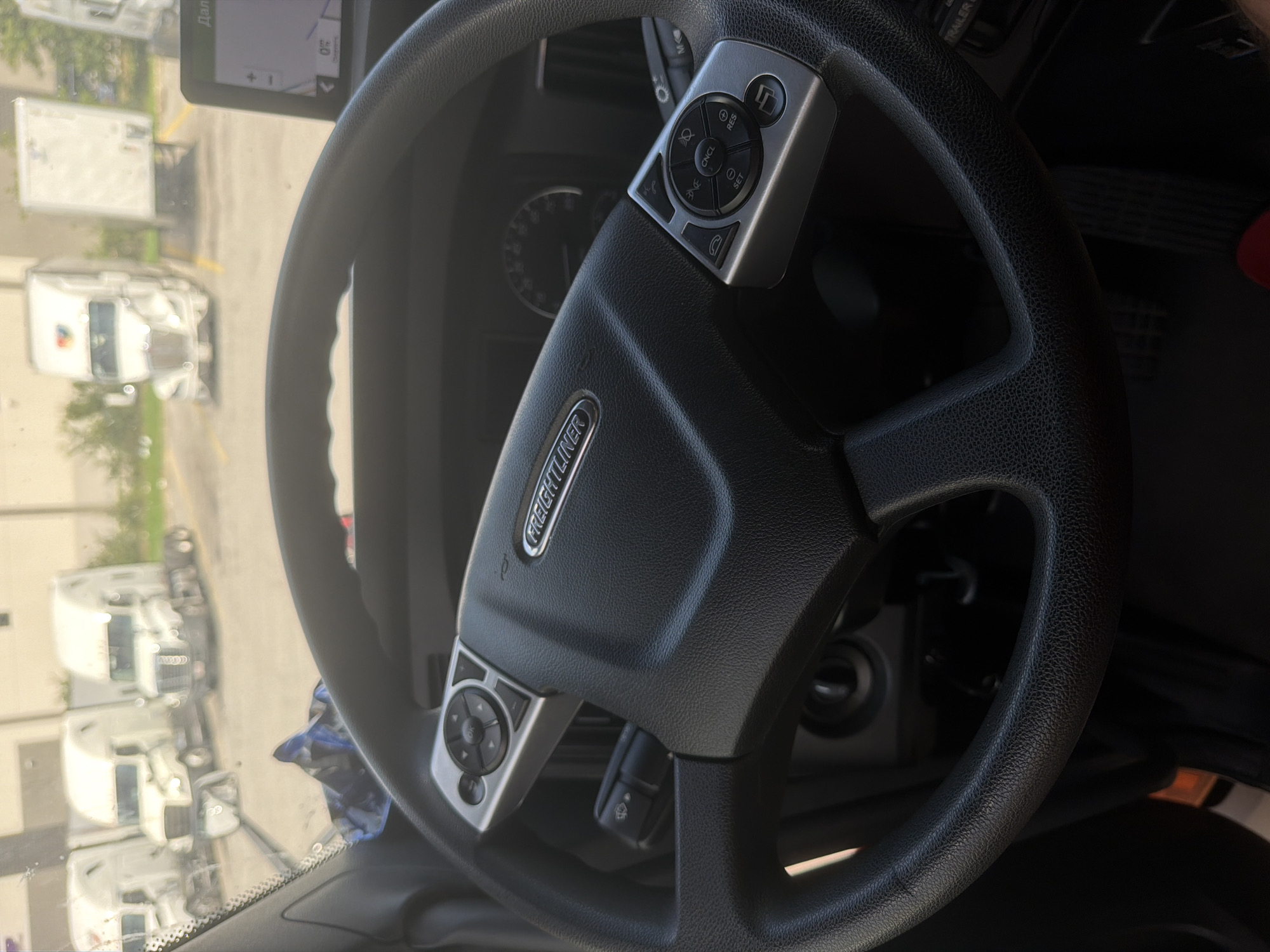 The image shows the interior of a Freightliner truck cab. The steering wheel is the primary focus, with its black textured leather and silver control buttons on either side. The Freightliner logo is visible on the center of the steering wheel. A GPS unit is mounted on the dashboard to the right, displaying a map. Outside the windshield, several white semi-trucks are parked in what appears to be a lot. One of the trucks has a colorful logo on its trailer. The background suggests an industrial or commercial area. The scene is brightly lit, likely during daylight. There are no people visible in the image.
